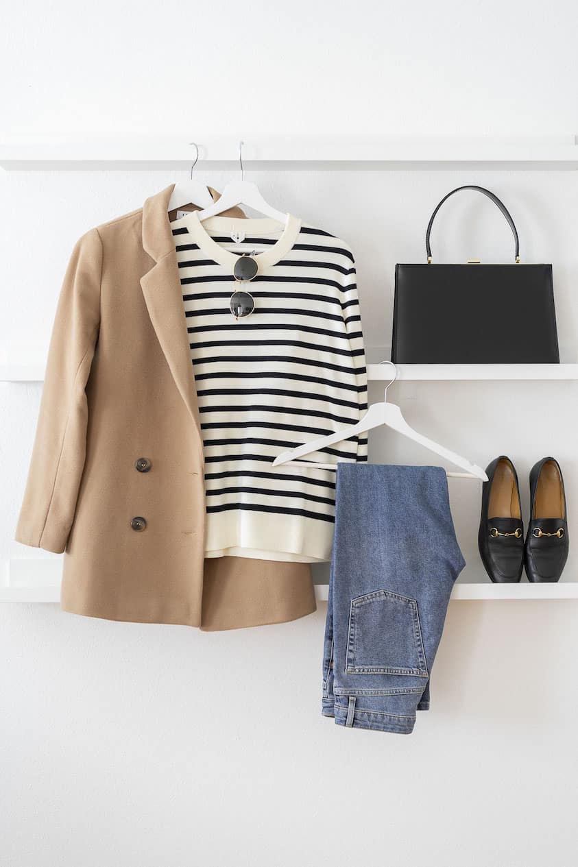 A tan blazer, black and white striped sweater, jeans, black loafers, black handbag, and gold sunglasses laid out on shelving.