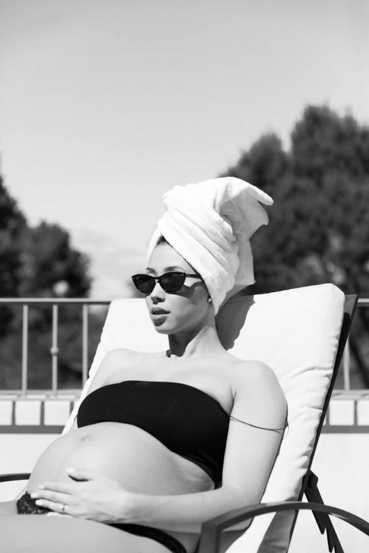 black and white image of a pregnant woman with a towel wrapped in her hair and oversized sunglasses sitting on a lounge chair in a black swimsuit