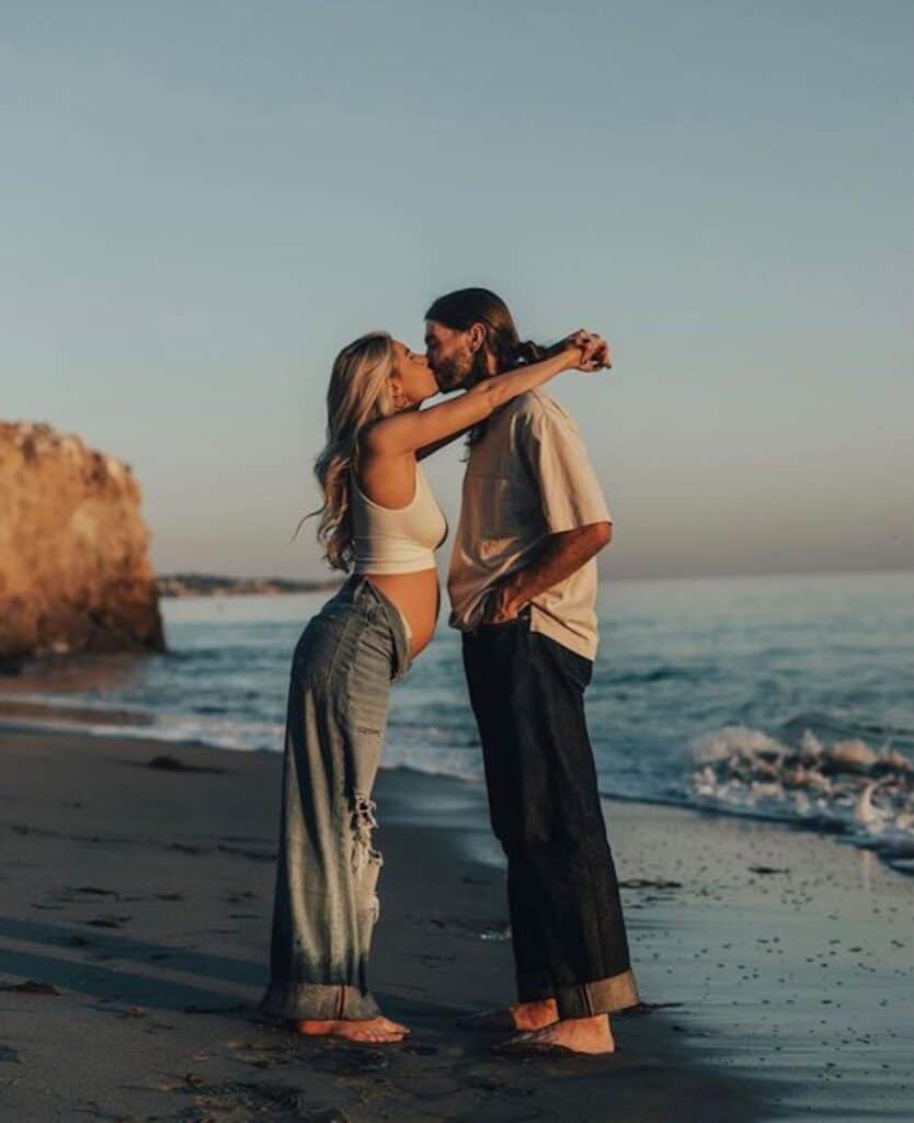 image of a man and woman standing on a beach at sunset, the woman is pregnant wearing rolled down jeans and a white tank top, leaning over to kiss the man