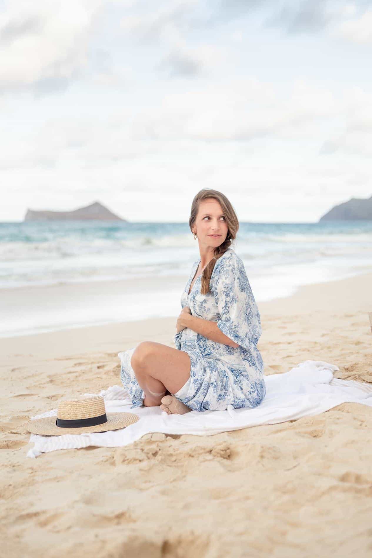 image of a woman sitting on a towel on the beach wearing a blue floral dress embracing her pregnant belly
