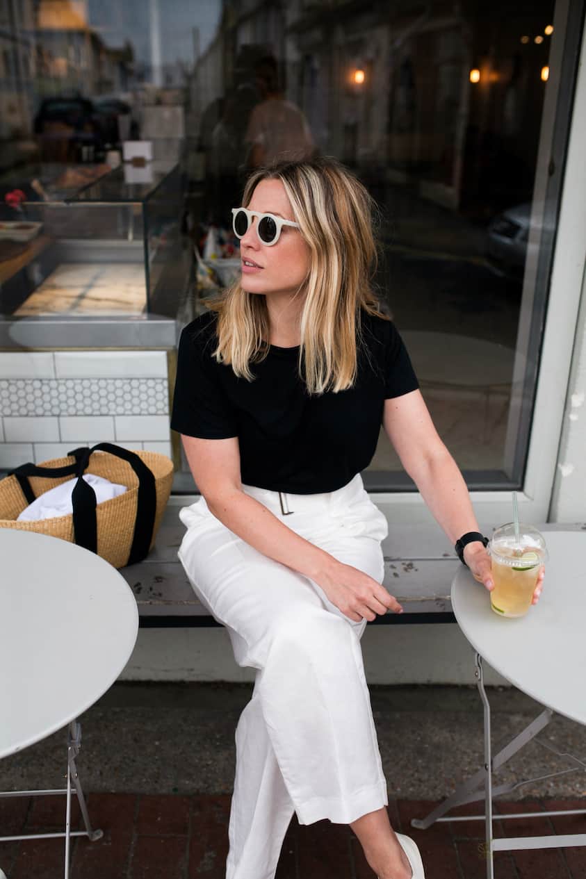 Woman wearing white linen pants, a black t-shirt and white sunglasses sips on a drink while sitting on a patio.
