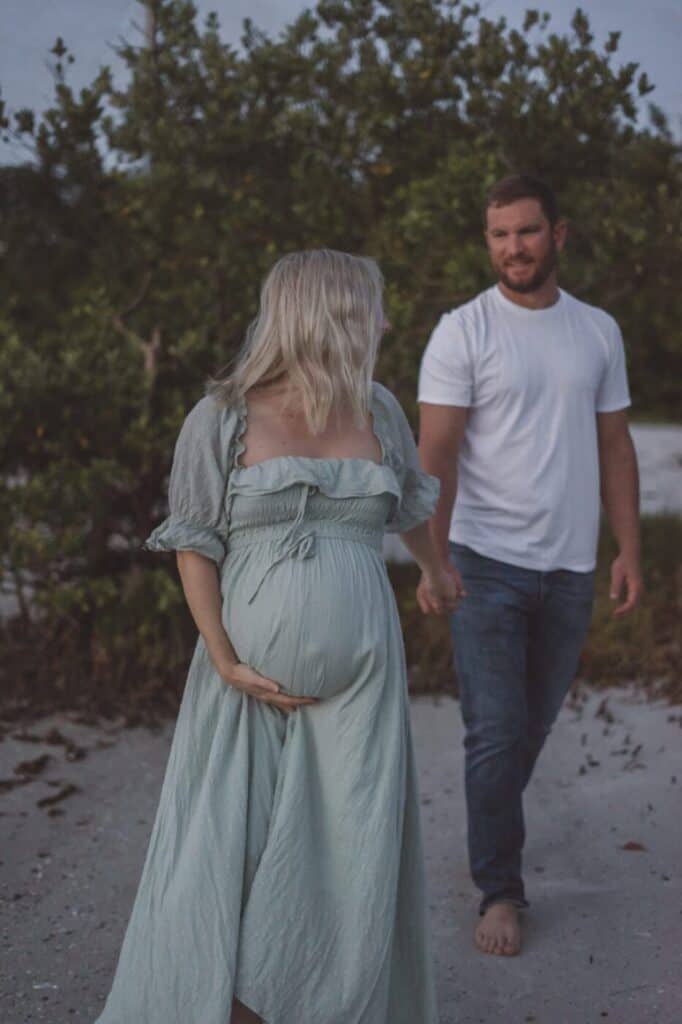 image of a pregnant woman leading a man onto a beach, she is wearing a long light green flowing dress and he is in a white t-shirt with jeans