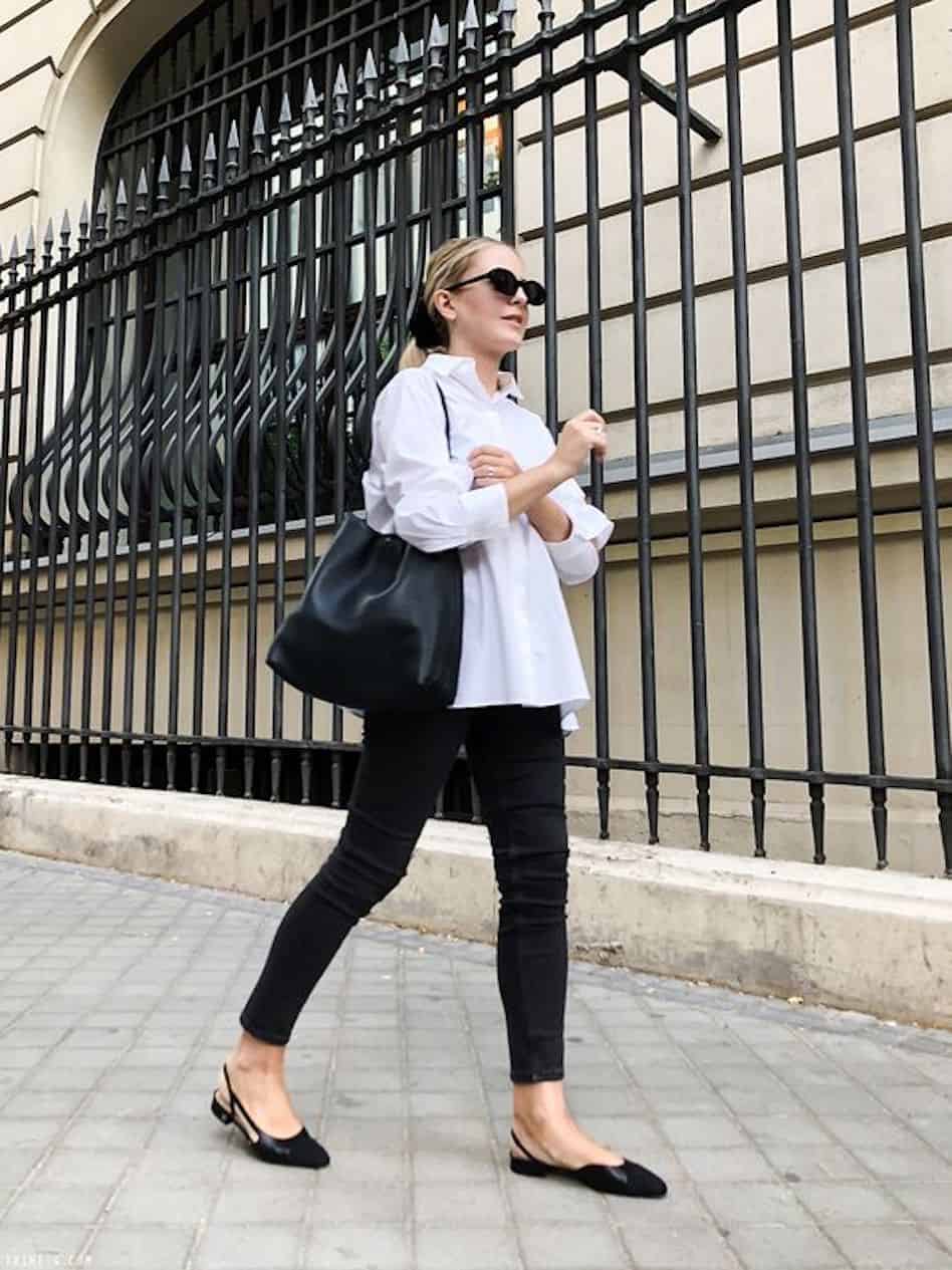 A woman wearing black jeans, black mules and a white button down walks in the city.