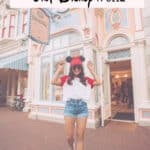 Girl at Disney wearing jean shorts, white sneakers, a white and red baseball tee, and a red Minnie Mouse hat.