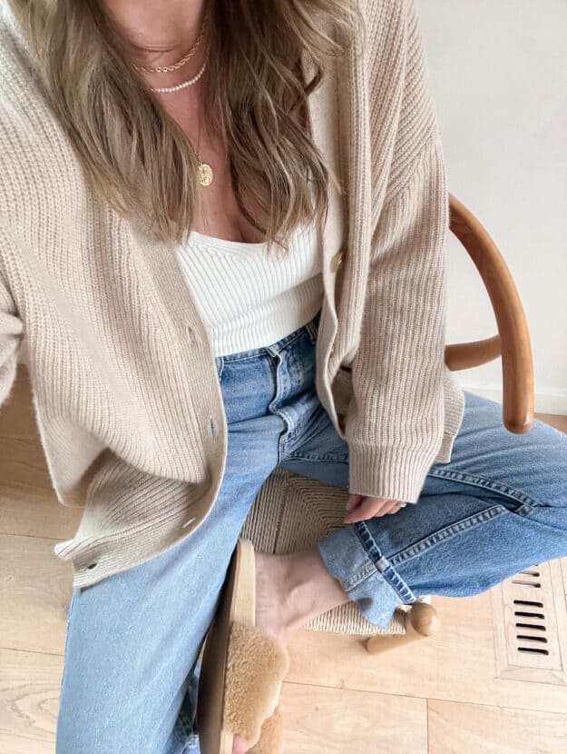 woman wearing a beige cashmere knit oversized cardigan from Jenni Kayne with an ivory ribbed tank top, jeans, and shearling slides