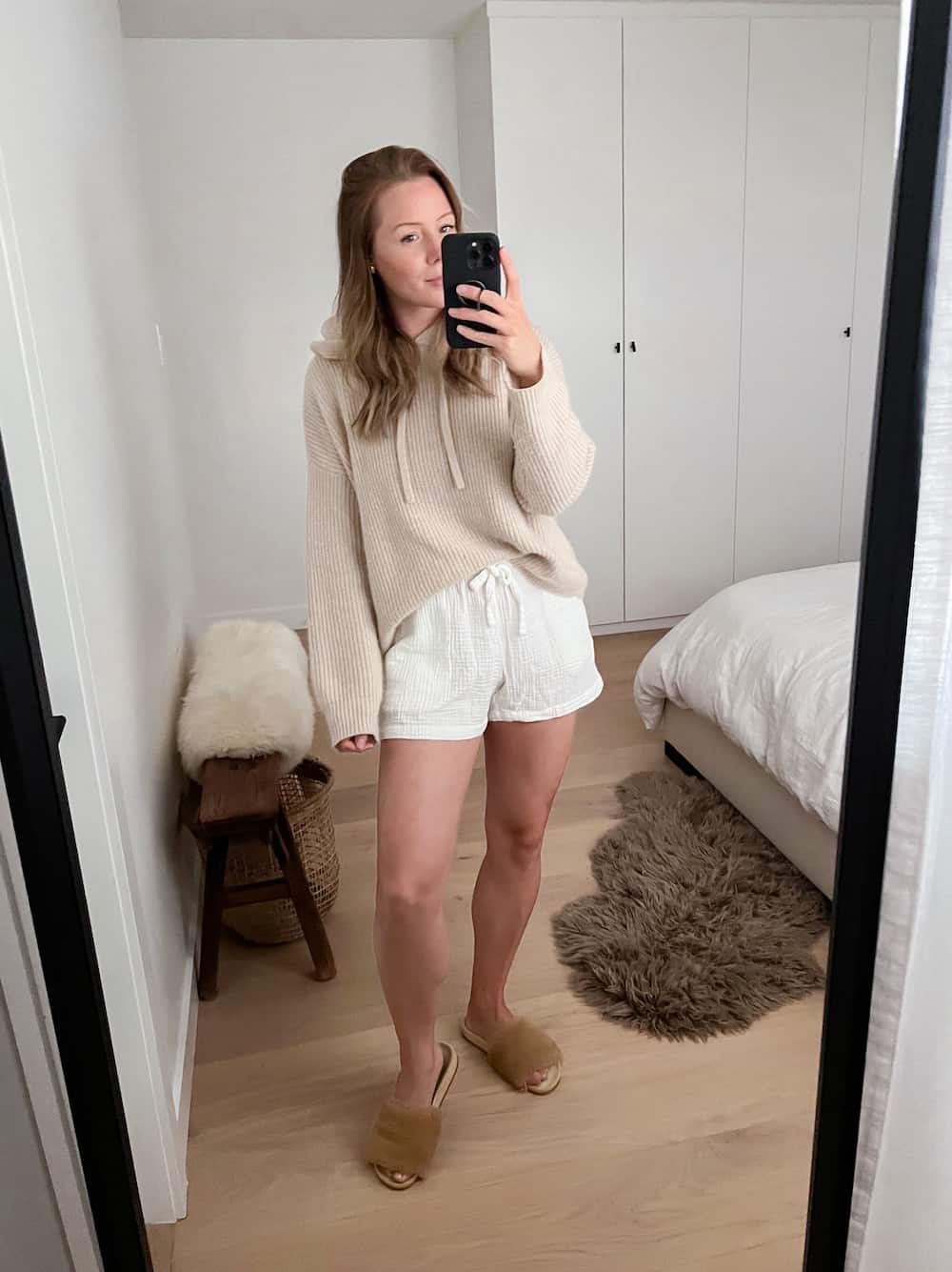 woman wearing a beige cashmere knit hoodie sweater from Jenni Kayne with white crinkle cotton shorts with shearling slides