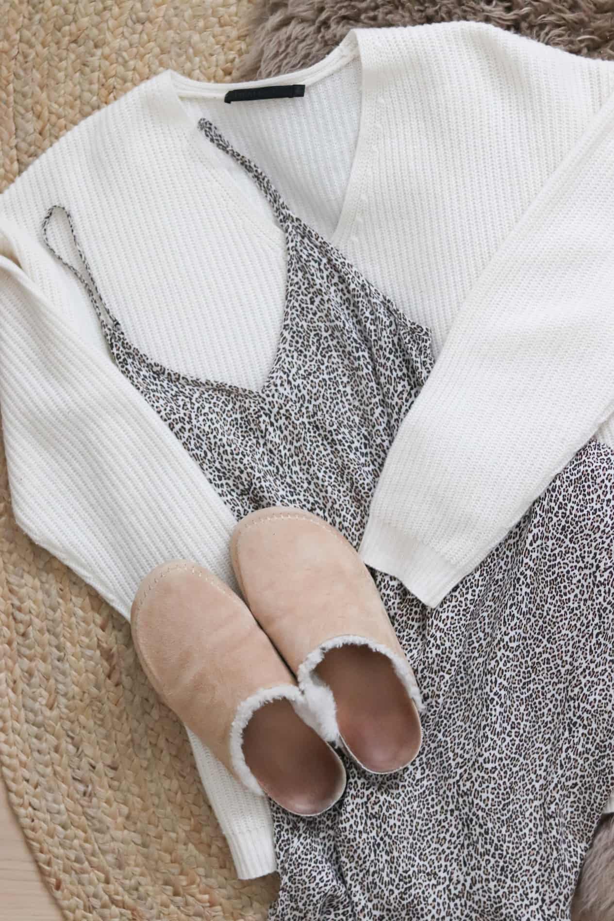 Flat lay of a white sweater, animal print spaghetti strap dress and cozy slippers.