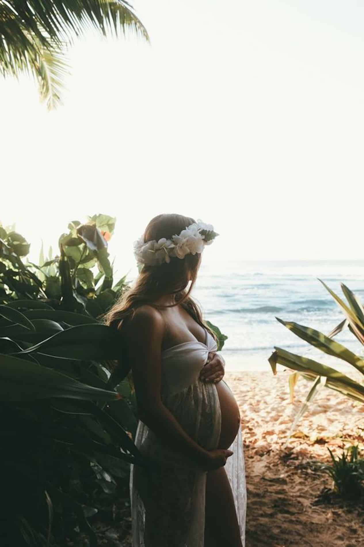 image of a pregnant woman standing by green bushes looking towards the beach and ocean with a white flowing gown and a flower crown in her hair