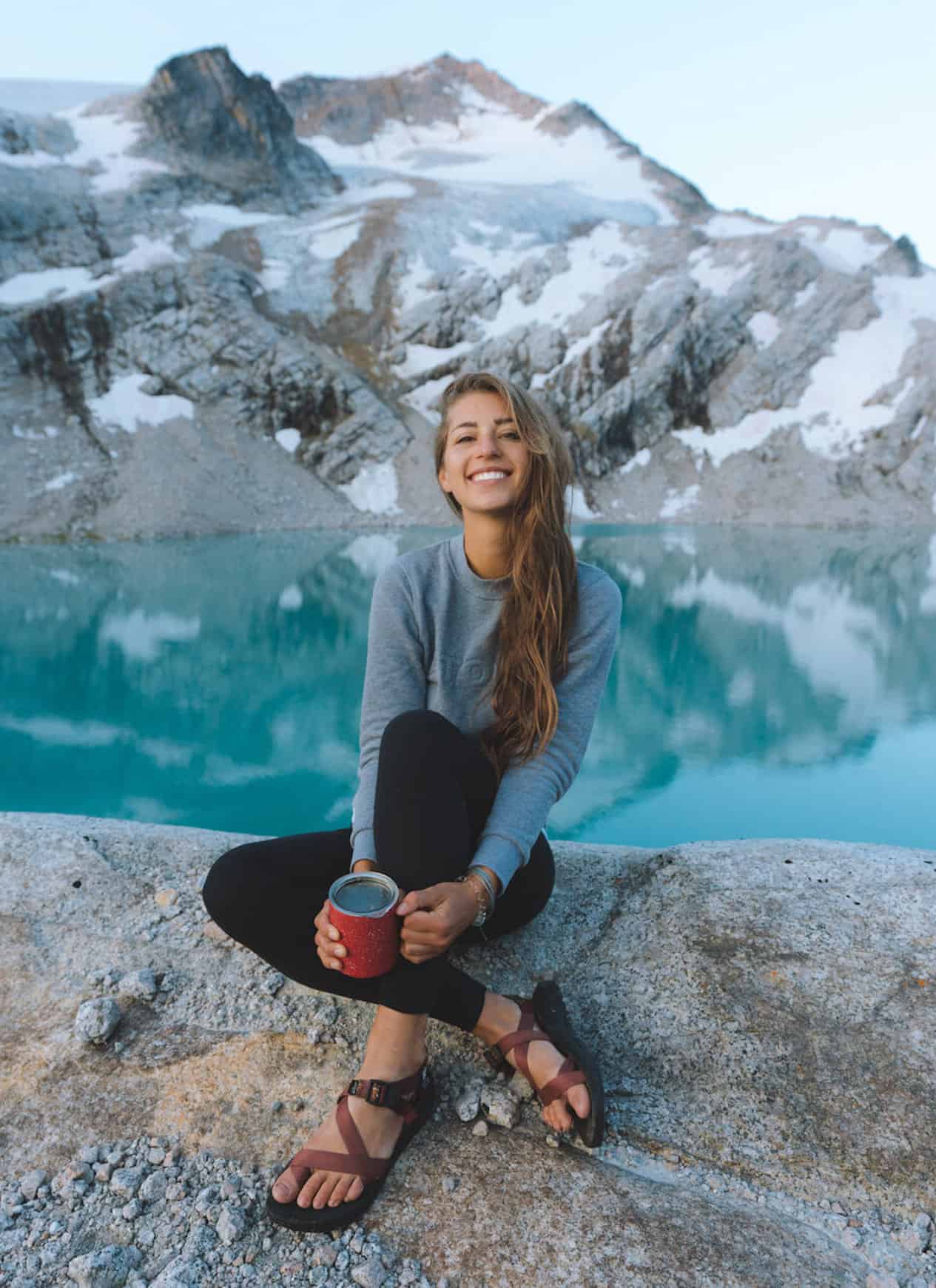 Girl sitting in the mountains near a lake wearing leggings, a sweatshirt and Chaco sandals.