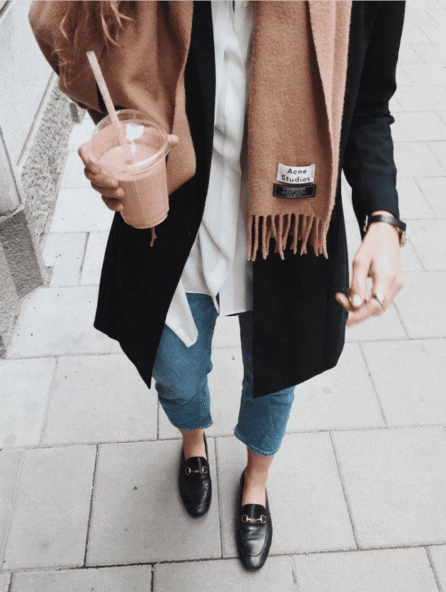 image of a woman from the shoulders down wearing a black coat, camel scarf, white shirt, jeans, and Gucci loafers, carrying a pink smoothie in her hand