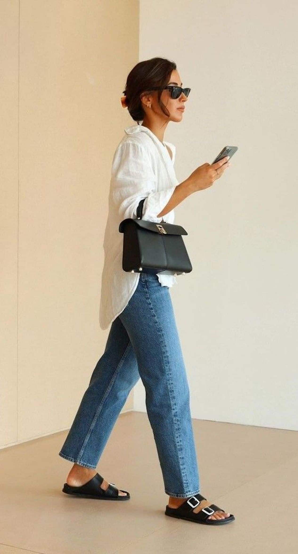 image of a woman standing from the side wearing an oversized white button up shirt, blue jeans, black sandals, and a black purse