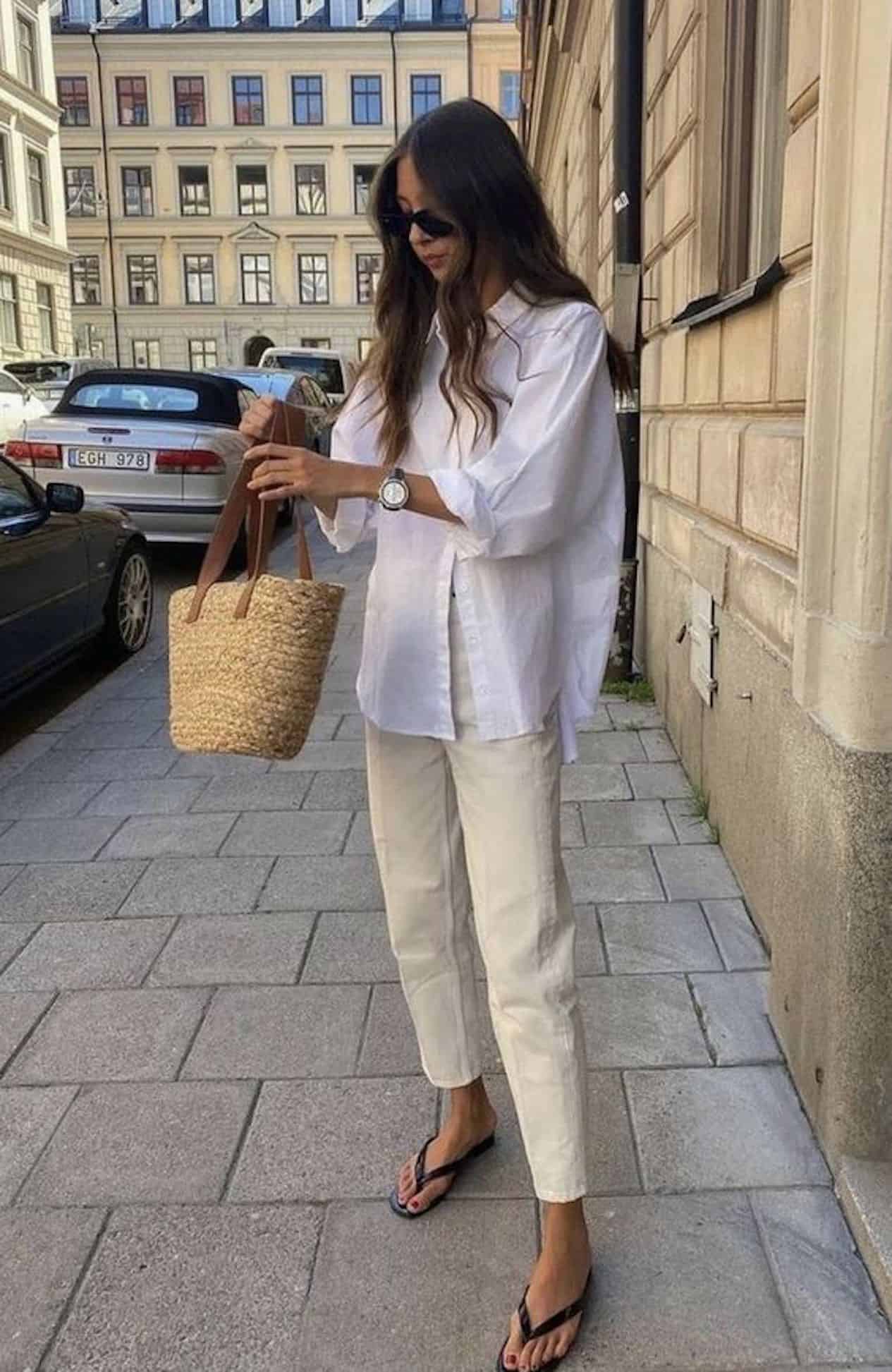 image of a woman standing on a sidewalk wearing a white button up shirt, cream pants, and black sandals, with a small straw bag
