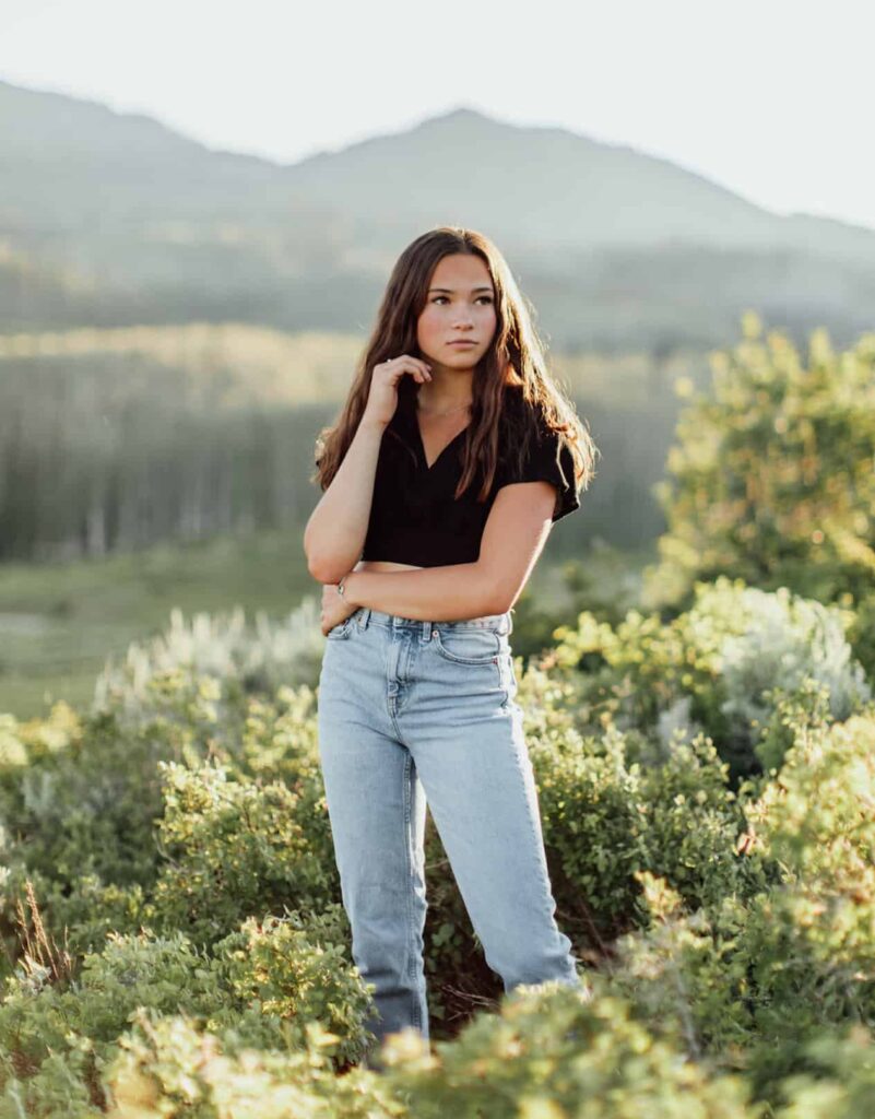 wholesome image of a teen girl with brown hair wearing a black t-shirt and jeans standing in a field with mountains in the background for senior photos