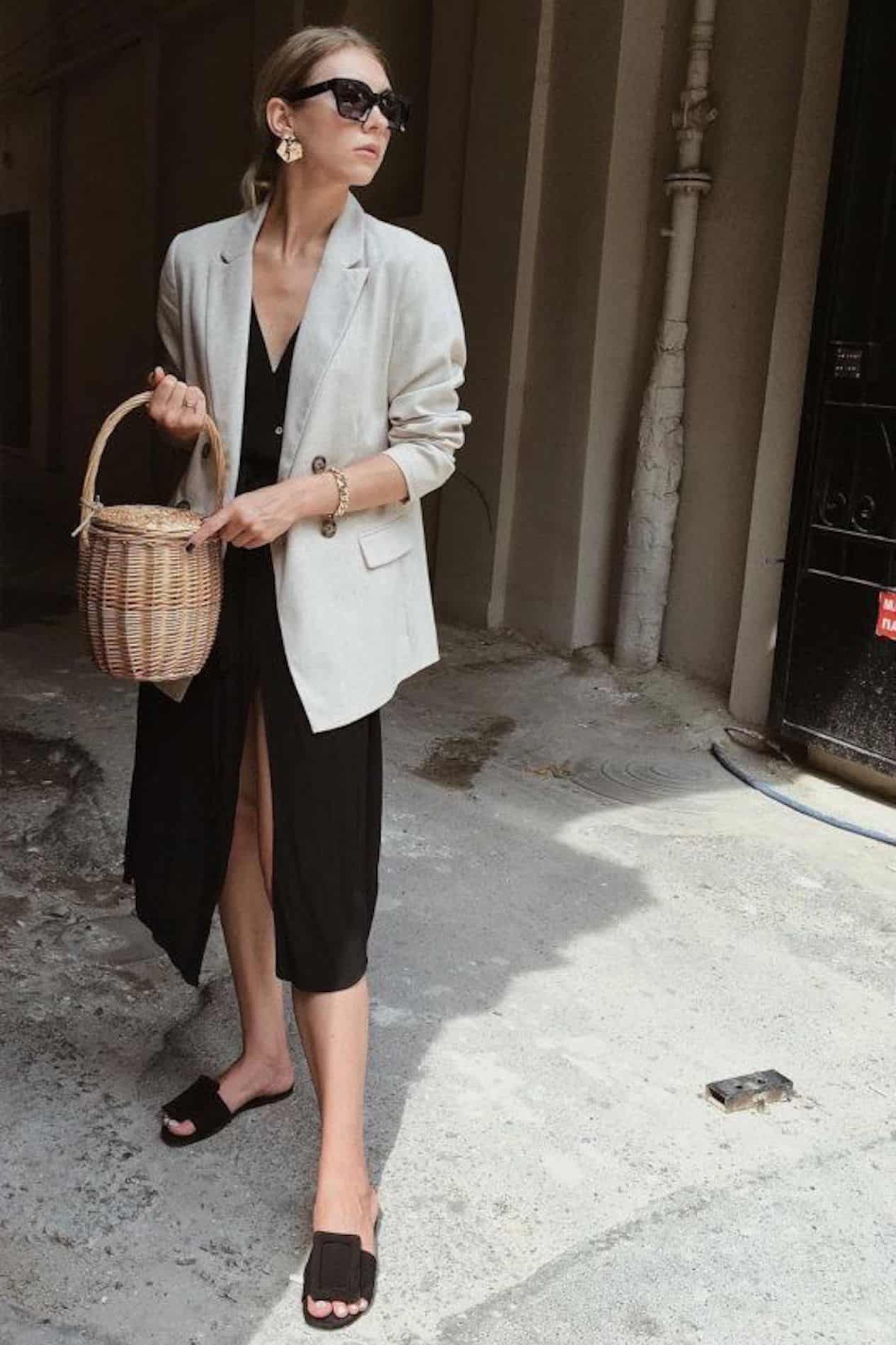 image of a woman with a black dress and linen blazer on holding a round basket bag and wearing black sandals