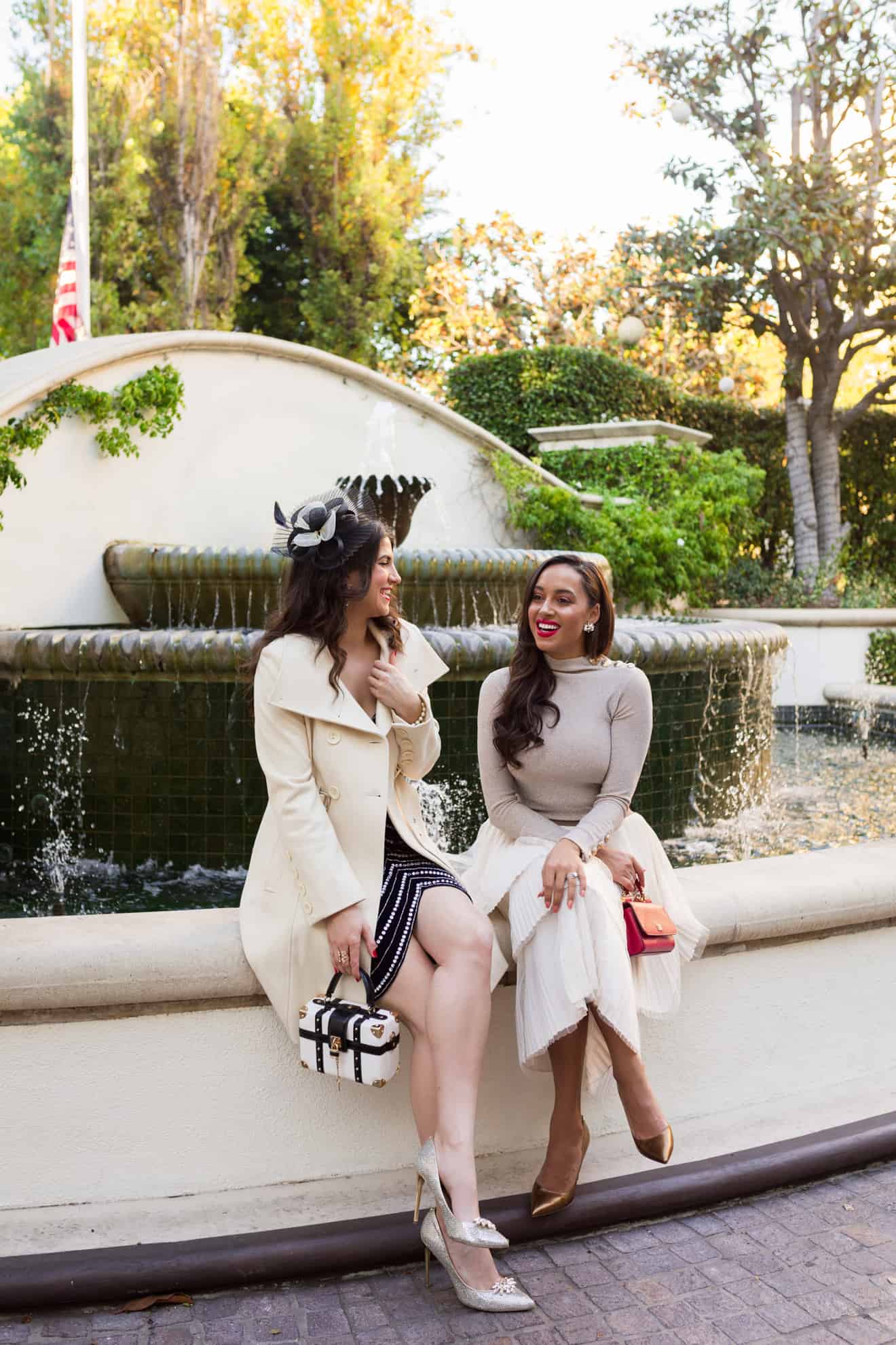 two women sitting on the edge of a fountain wearing formal clothing, including a dress and overcoat, tulle skirt, and heels