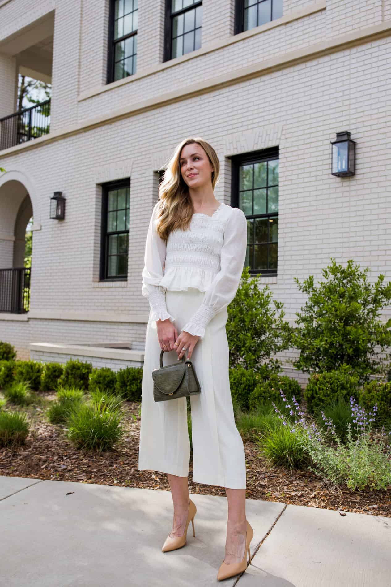 a woman standing on a sidewalk with greenery in the background wearing a white blouse and wide leg cream trousers and heels 