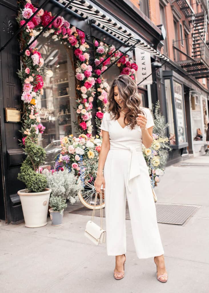 image of a woman standing on a sidewalk with a floral display in the background wearing a white dressy jumpsuit and heels