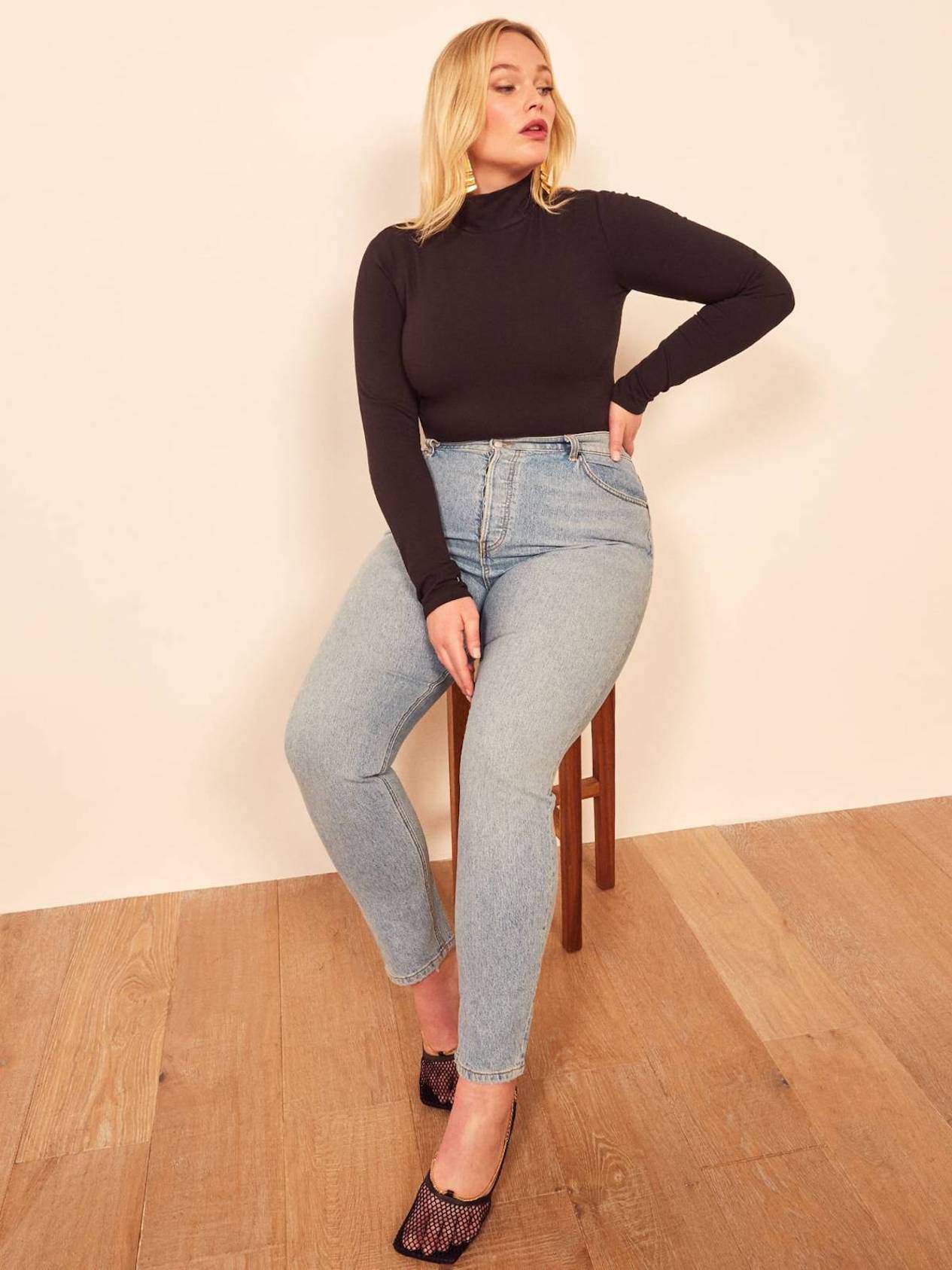 Woman sitting in a chair wearing light wash jeans, and a black mock-neck top.