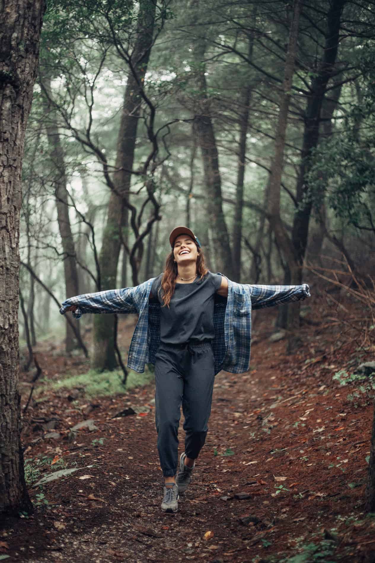 Girl walking in the forest wearing a black joggers and a black t-shirt and a blue flannel.
