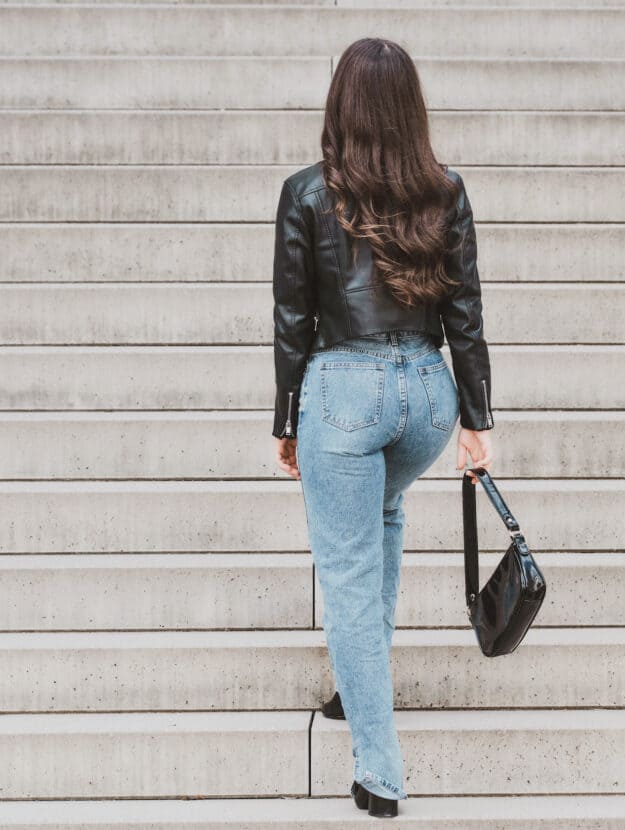 Woman walking up a flight of stairs wearing light wash jeans and a leather jacket.
