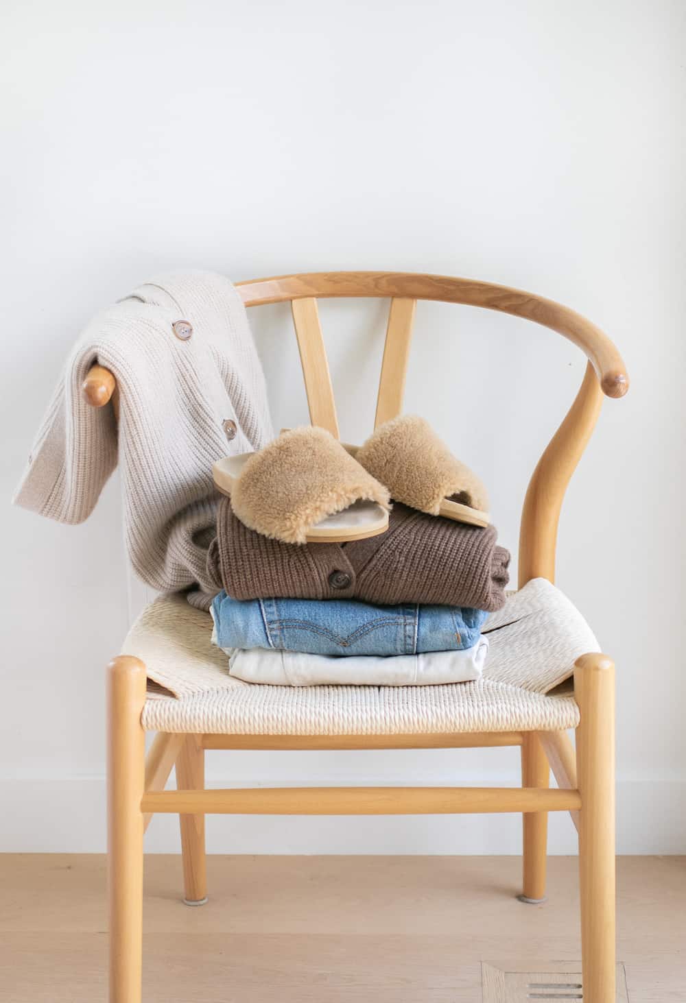 a stack of clothing on a wishbone chair including two Jenni Kayne cocoon cardigans, jeans, and a pair of shearling mules