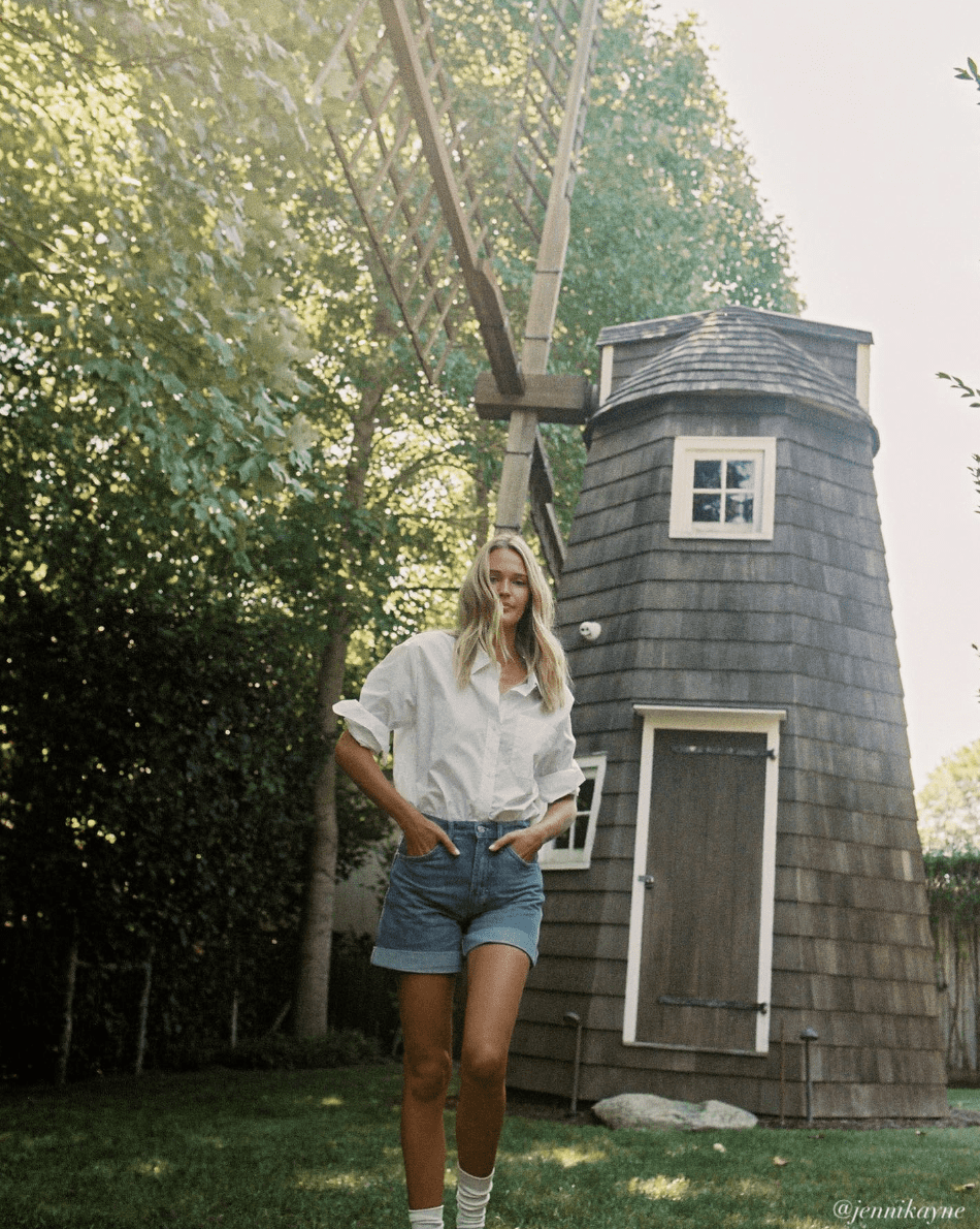 woman in front of an old windmill wearing a summer outfit with a white button up shirt and denim shorts