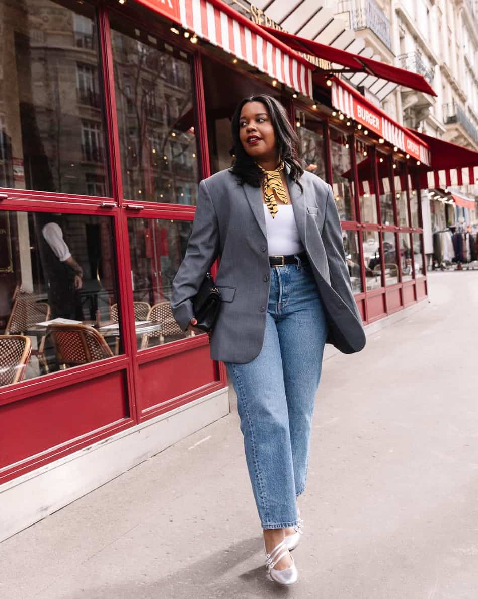 Black mid-size curvy woman wearing a grey blazer over a white top with straight leg jeans and silver Mary janes