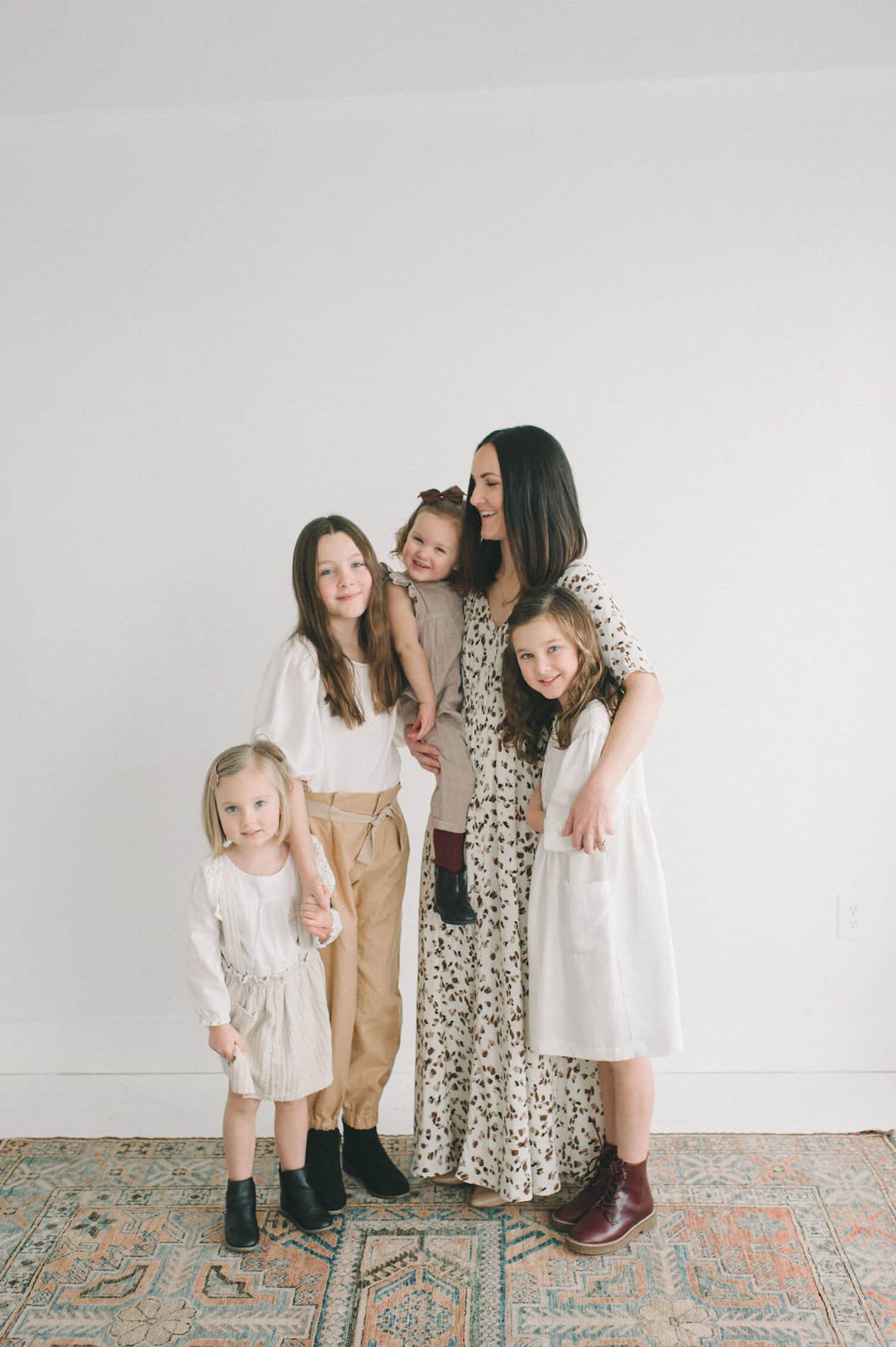 image of a mother and her four daughter standing in a white room on a vintage carpet wearing neutral clothes for a photoshoot