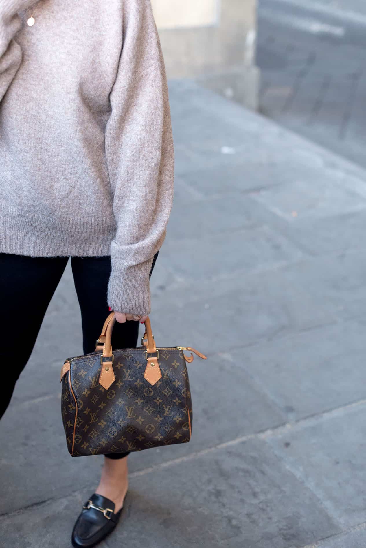 image of the side of a woman's body wearing black pants, a grey sweater, black loafers, and holding a Louis Vuitton bag in one hand