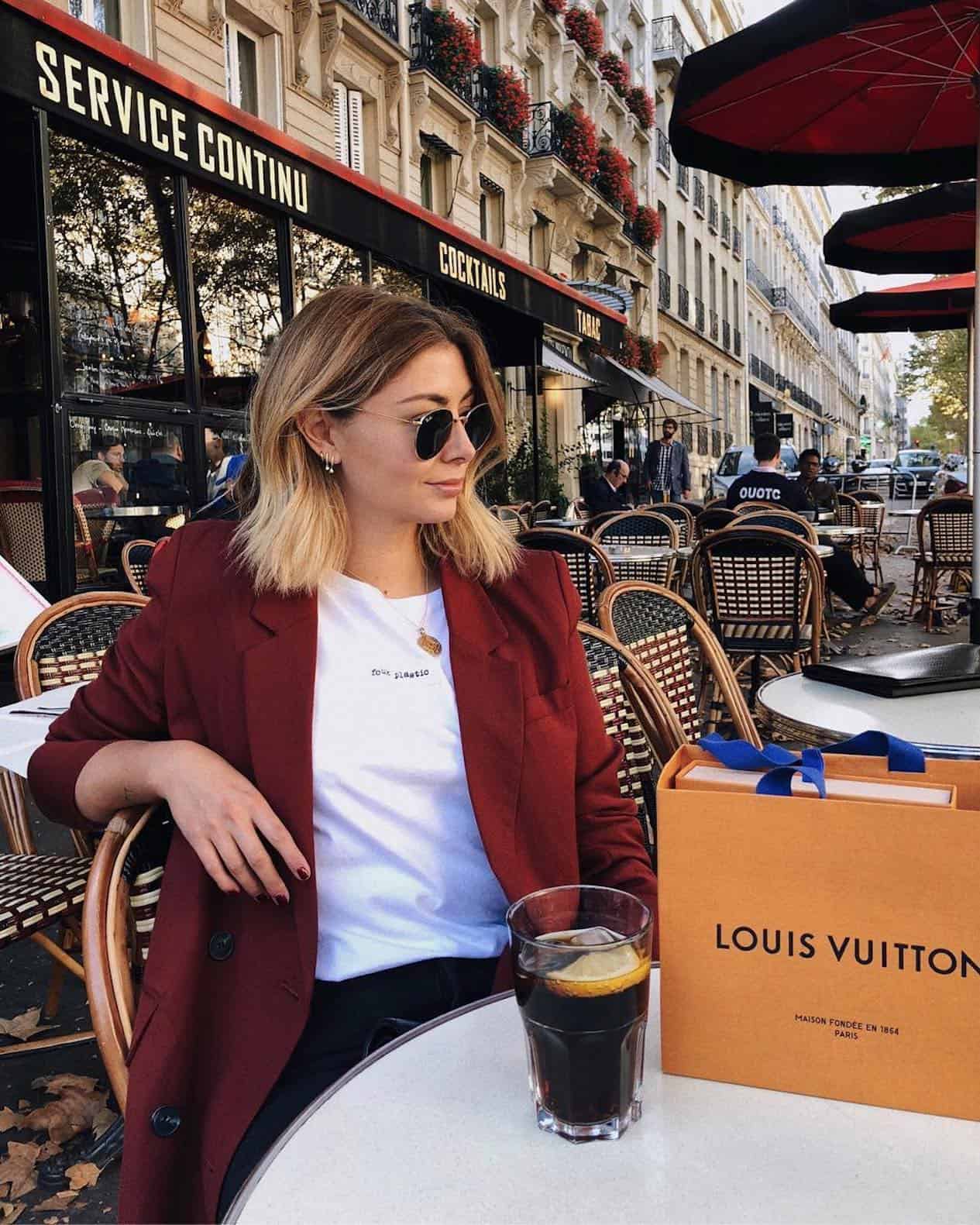 a woman sits on a chair in a cafe with a red blazer and white t-shirt, sitting on the table is a Louis Vuitton bag