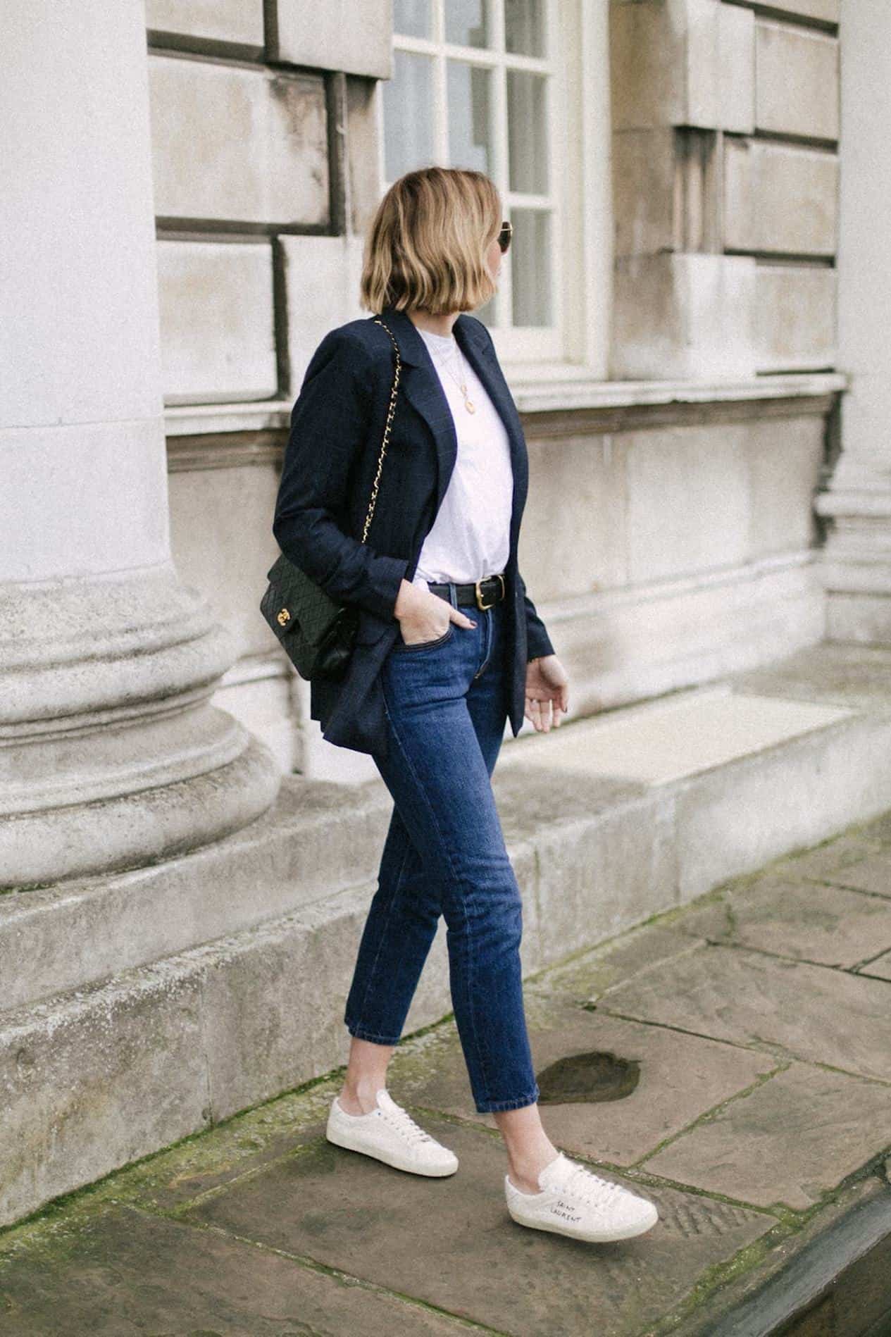 image of a woman standing on a sidewalk wearing blue jeans, a white t-shirt, a navy blazer, and white sneakers