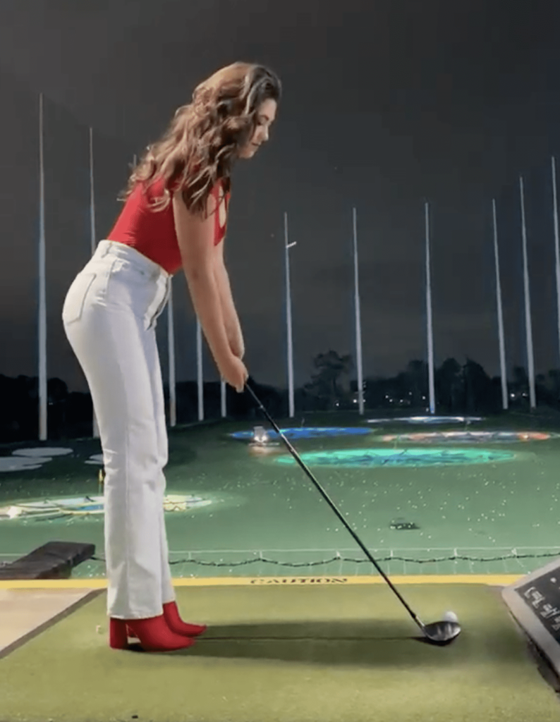 image of a woman with brown long hair playing golf at Topgolf wearing a red top, white jeans, and red heeled boots
