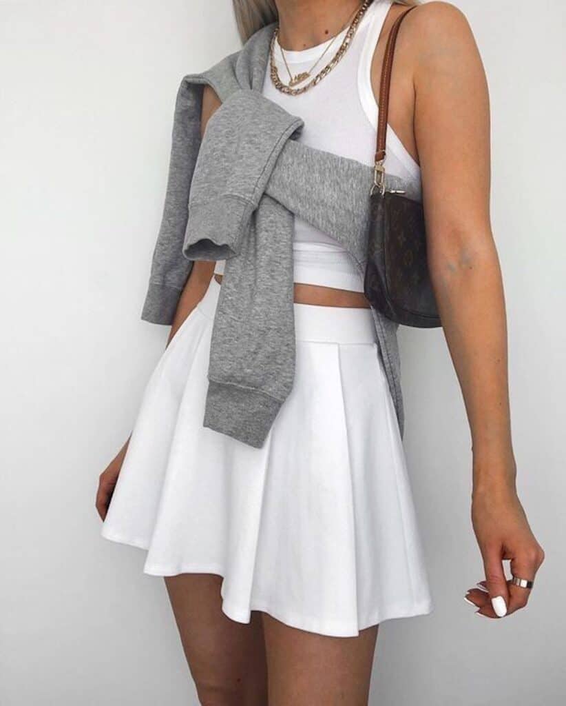 image of a woman from the shoulders down wearing a white crop top, white golf skirt, with a grey sweatshirt wrapped around her body and a Louis Vuitton purse