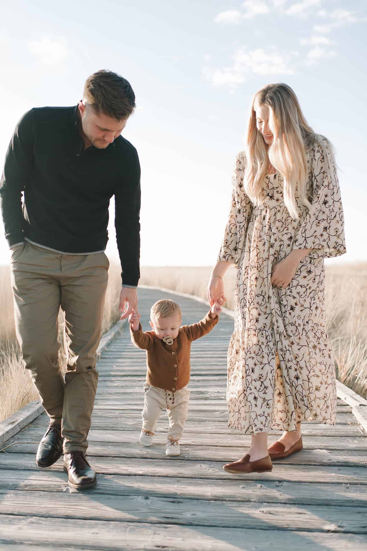 image of a family of three walking down a coastal boardwalk, the mom in a floral maxi dress, the dad in chino pants and a sweater, and the baby in a brown sweater and tan pants