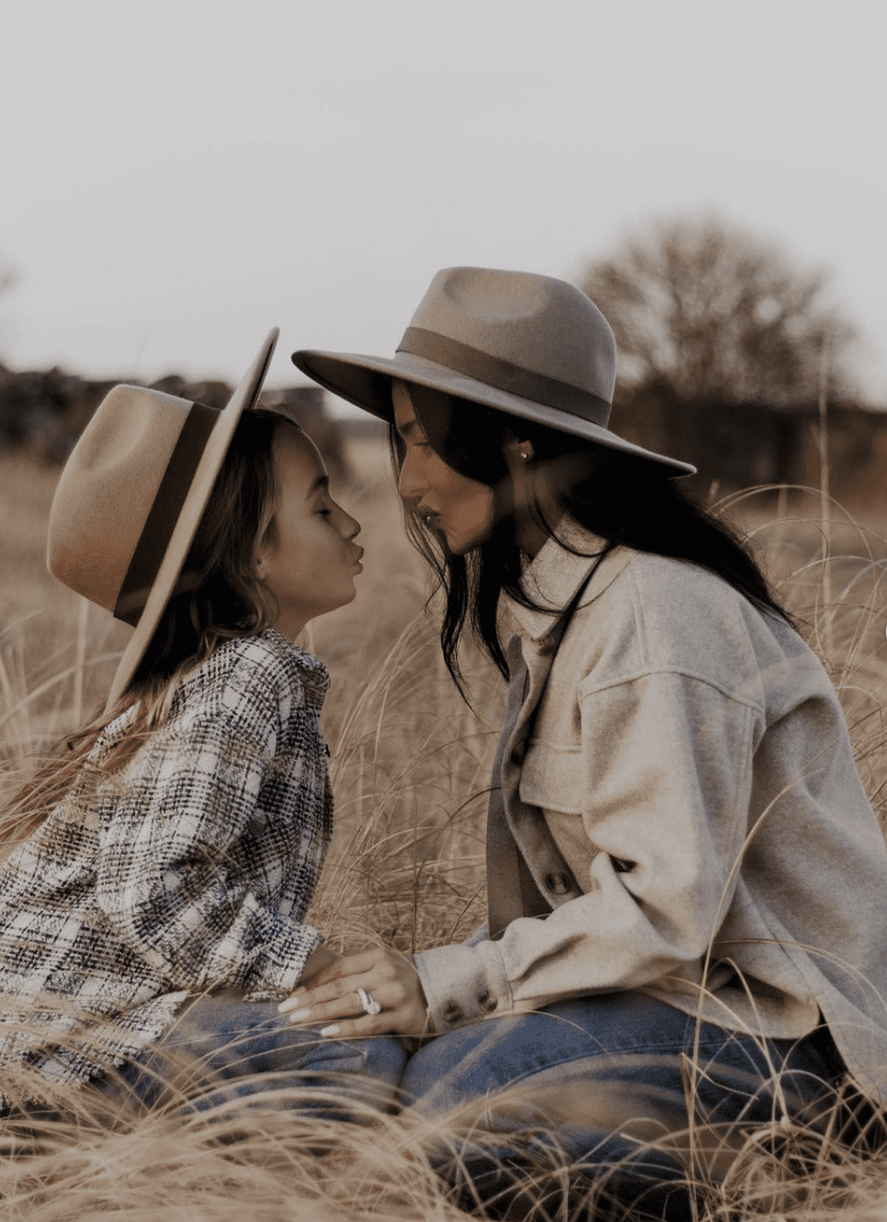 image of a woman and her daughter in a grassy field wearing wool fedora hats, shirt jackets, and jeans, looking at each other lovingly