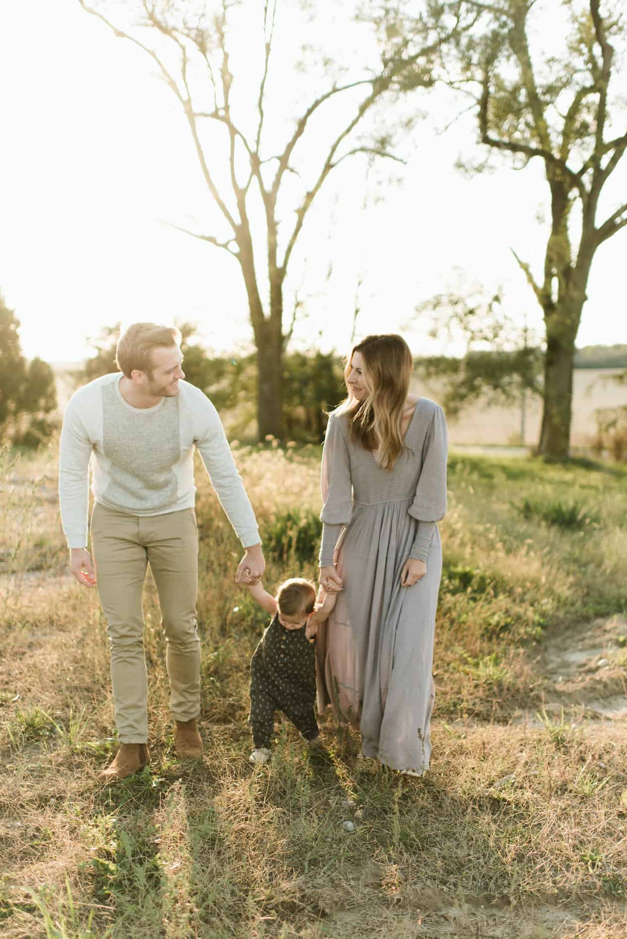 image of a family of three walking through a countryside meadow at sunset, the mom wearing a long mauve dress, the baby in a green romper, and dad in green pants and an ivory sweater