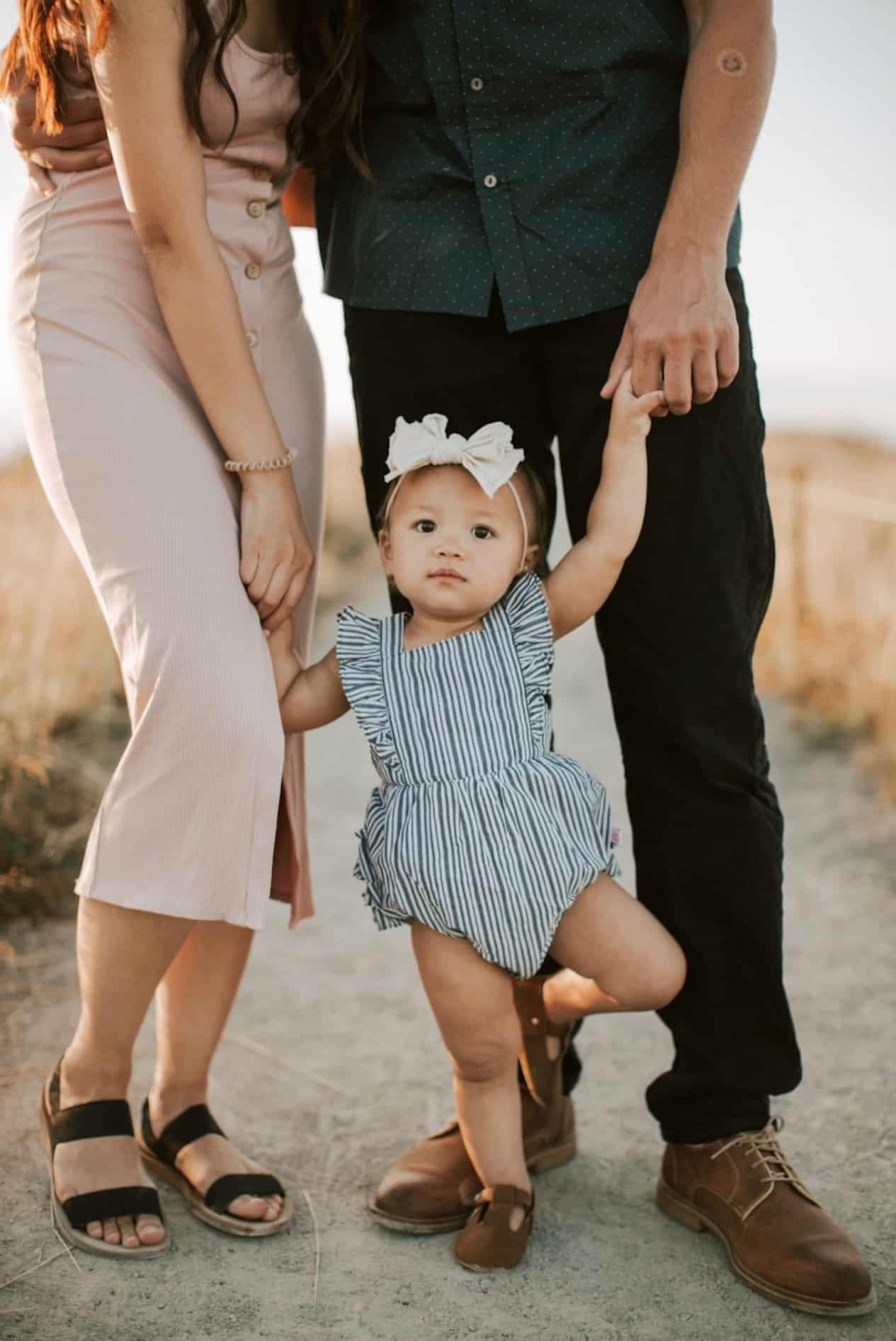 image of a little girl in a blue striped romper holding her mom and dads hands for a family photoshoot