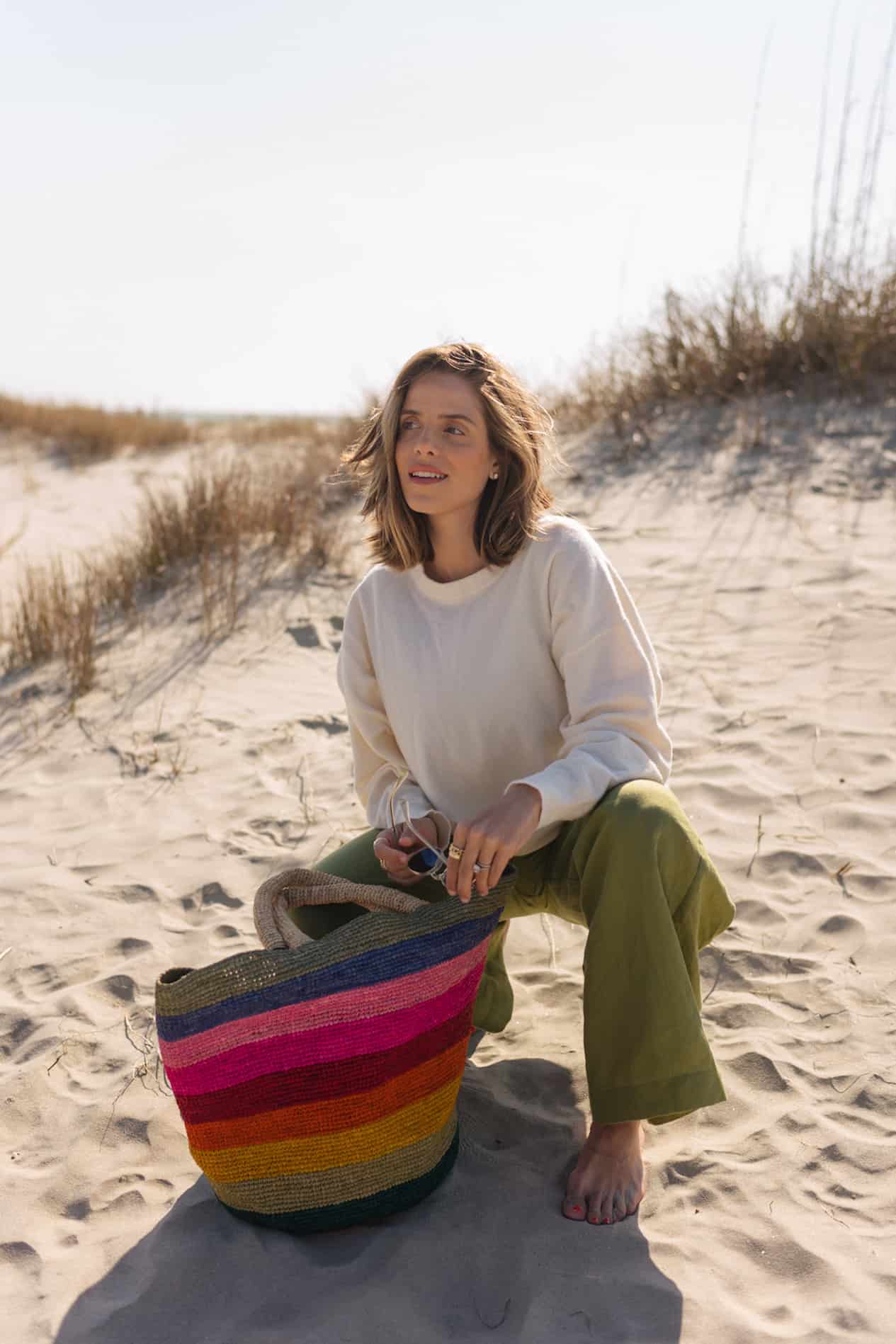 image of a woman wearing green wide leg linen pants, an ivory sweater, and a rainbow striped straw bag, bending down on a beach
