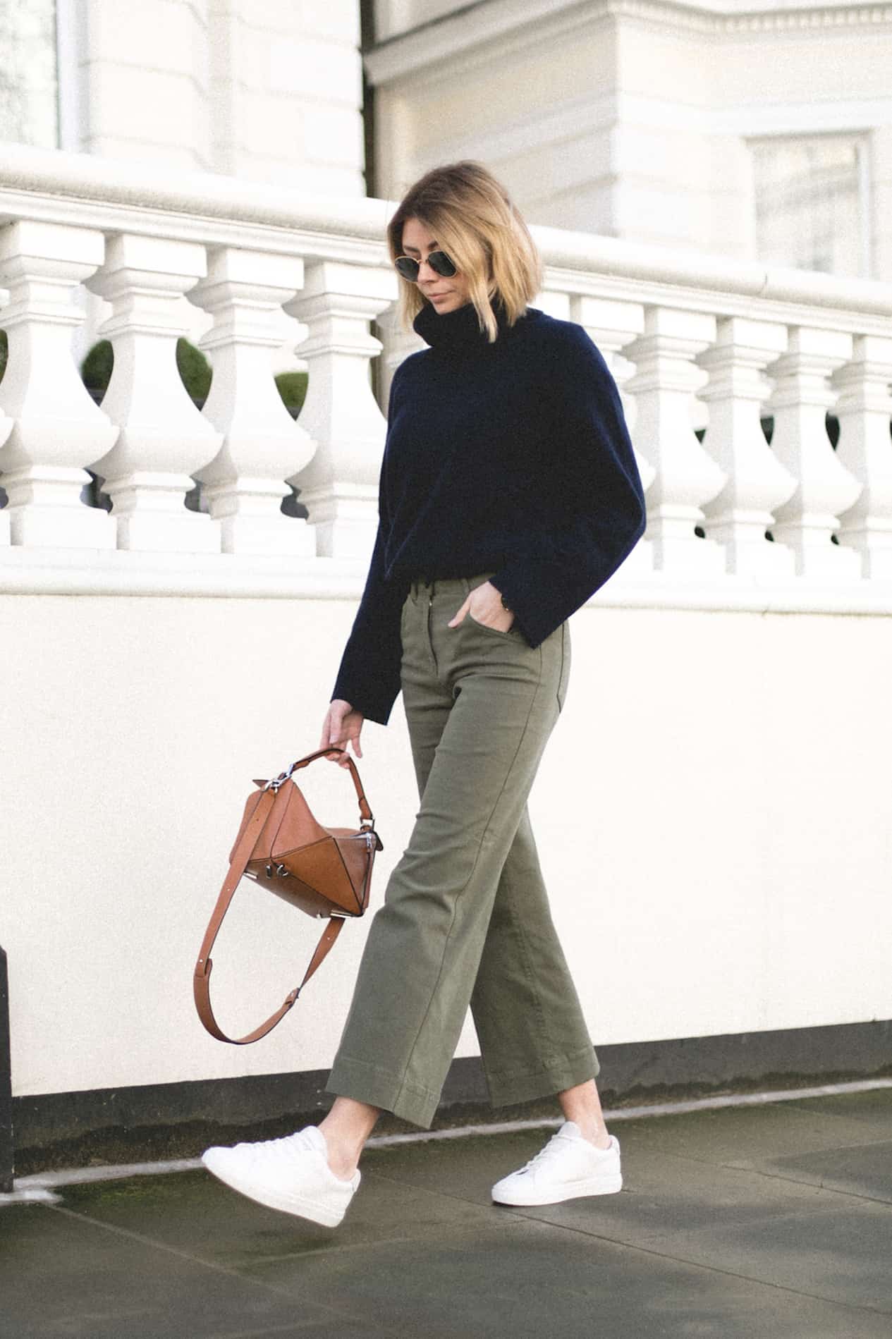 image of a woman walking down the sidewalk wearing green pants, a navy sweater, and white sneakers, holding a designer brown purse