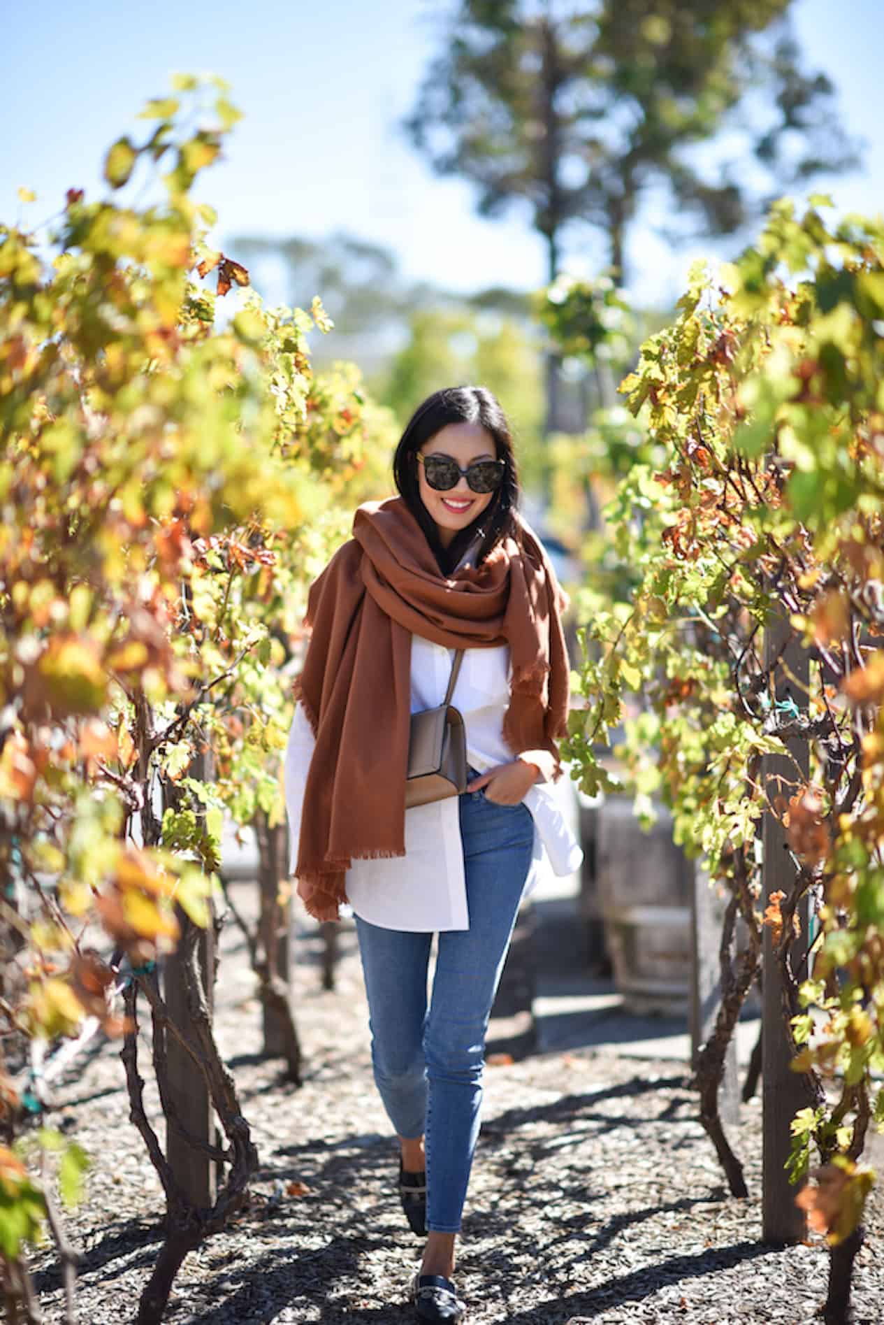 image of a woman walking through a vineyard wearing a white button up shirt, jeans, loafers, and a large brown scarf