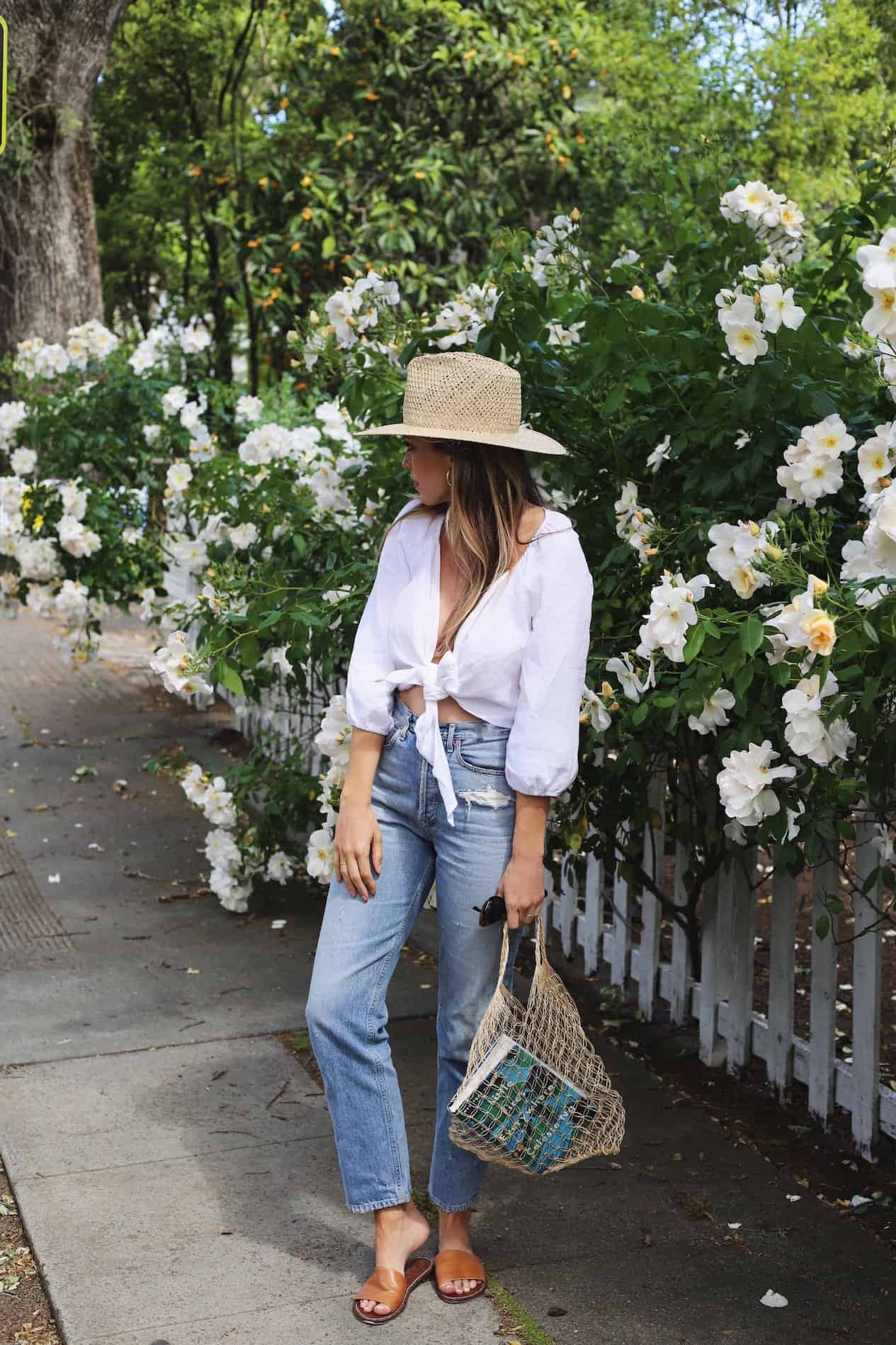 image of a woman standing on a sidewalk in front of flower bushes wearing a white blouse, blue jeans, and a woven hat