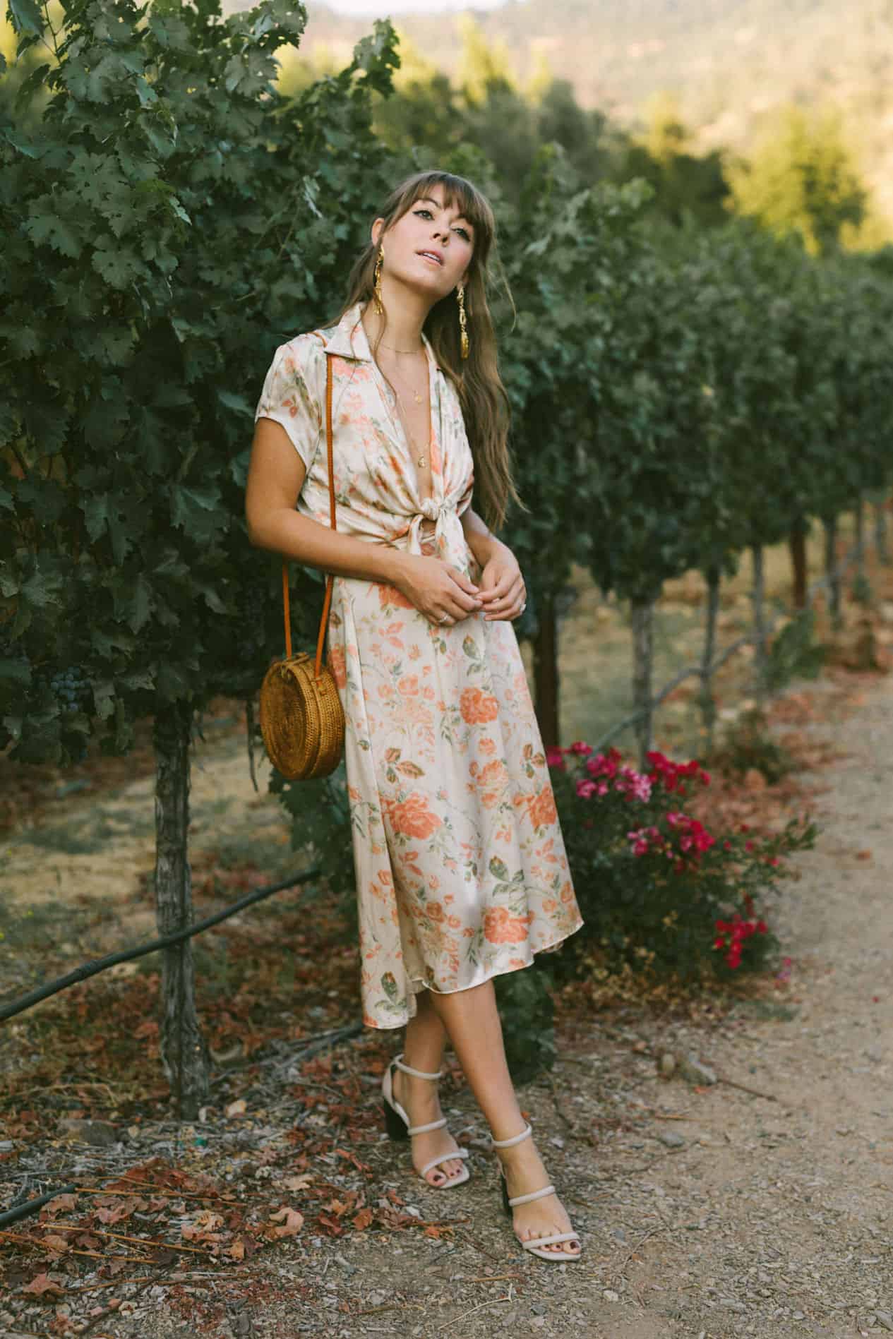 image of a woman wearing a silk floral dress with a straw round bag standing in a vineyard along grape vines