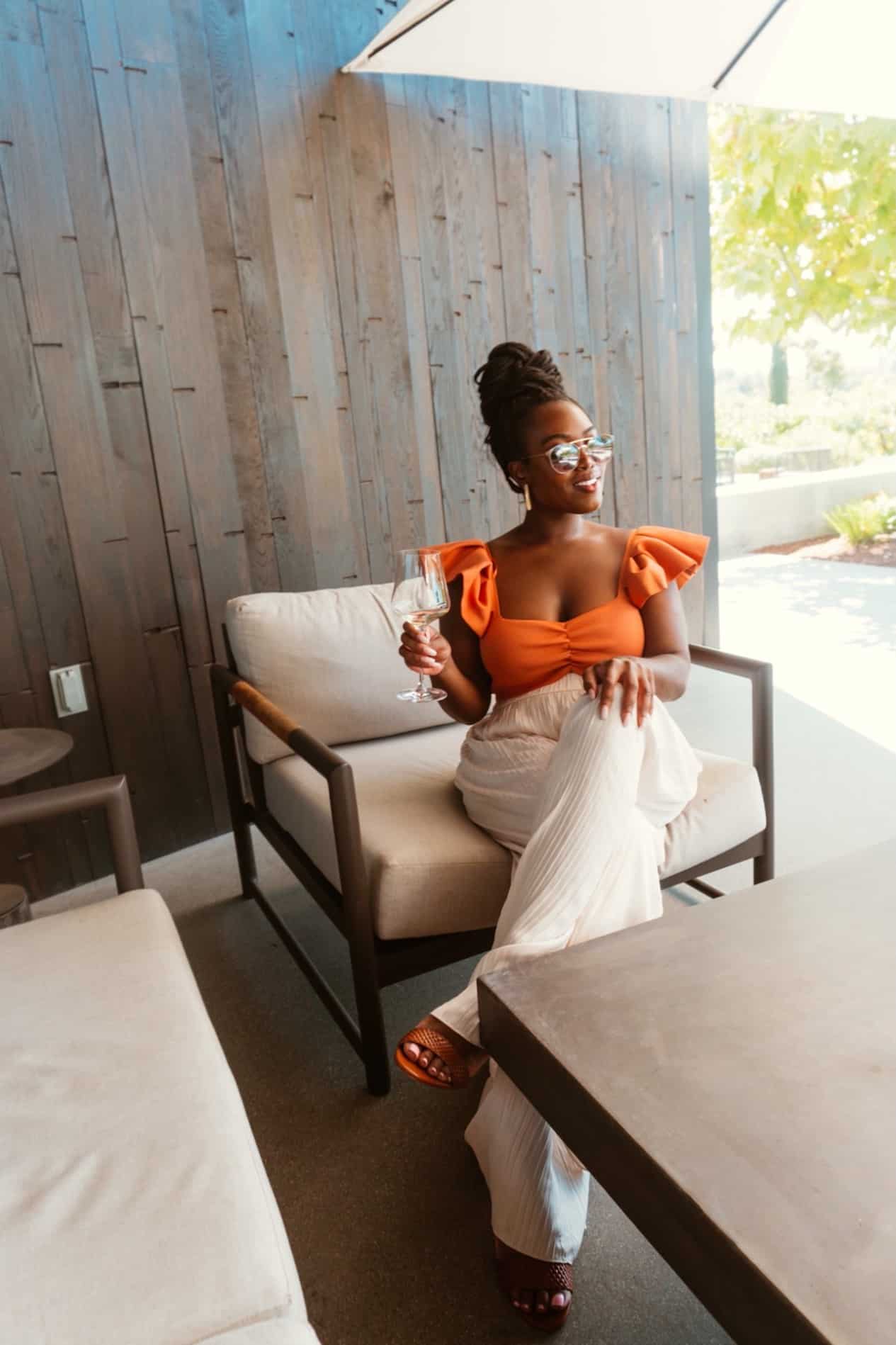 image of a black woman sitting on a large chair outdoors with a wine glass in her hand wearing an orange blouse and white linen pants