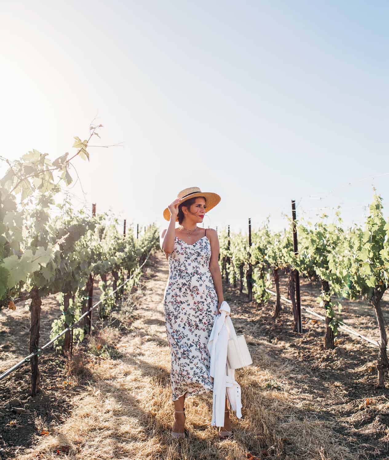 image of a woman standing in a winery in the vineyard wearing a floral midi dress and straw hat