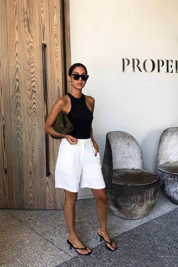 image of a woman standing in front of a large door wearing white Bermuda shorts, a black tank top and black kitten heel sandals