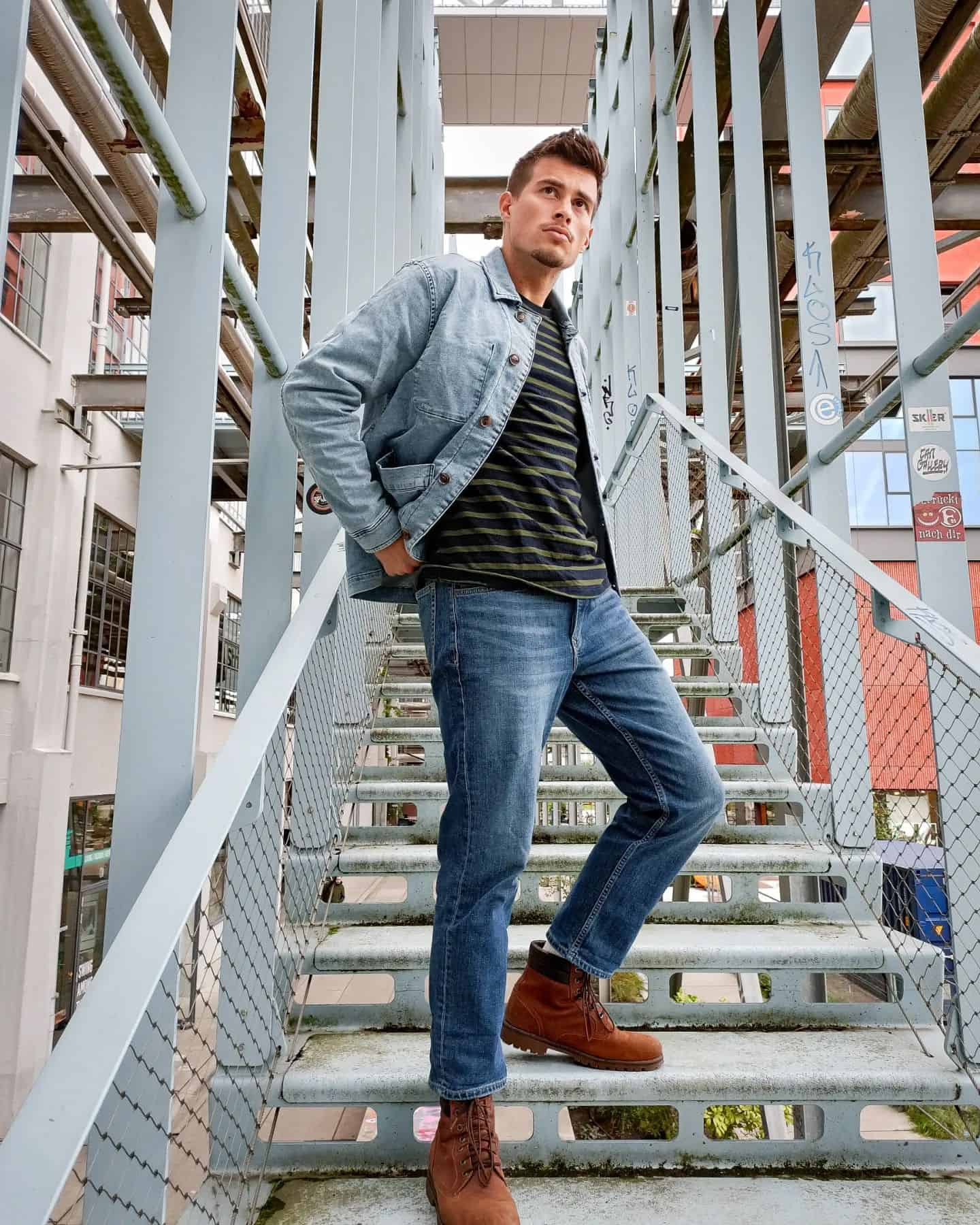 man standing on a stairwell wearing a denim jacket, dark shirt, bootcut jeans, and brown boots