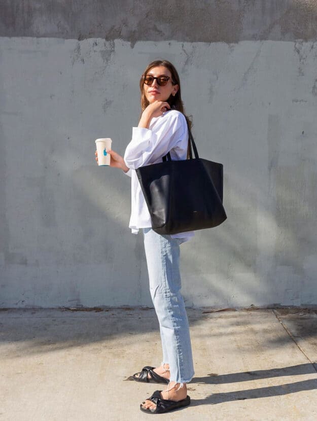 Woman wearing a white button down shirt with a black leather tote bag, jeans, and black sandals