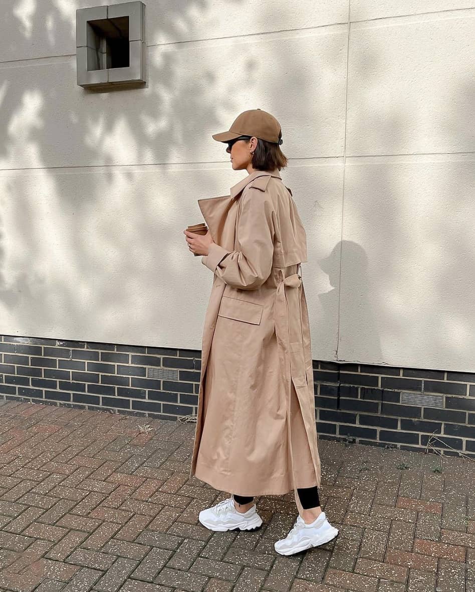 Woman wearing a beige ball cap with a long tan trench coat, black pants, and sneakers