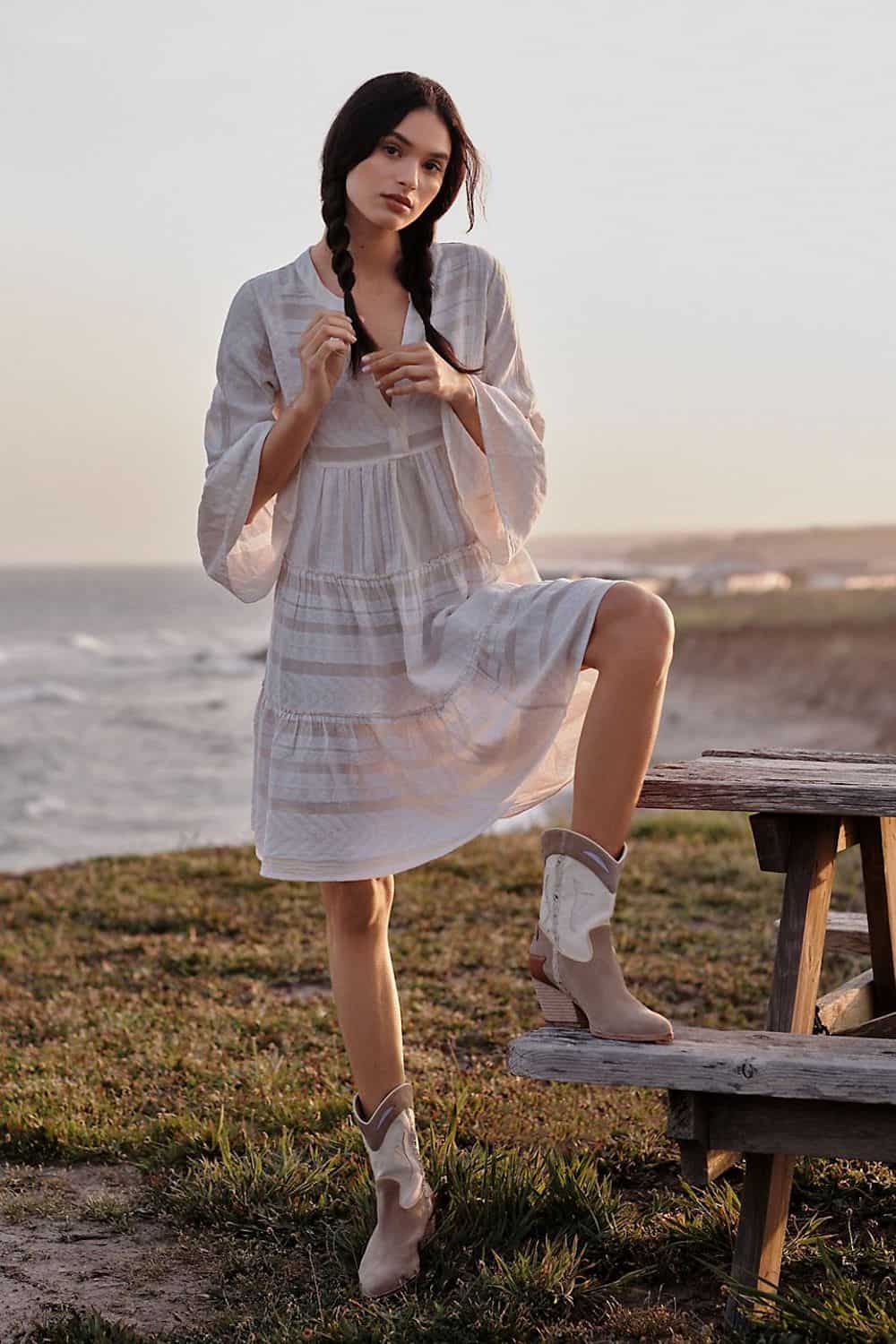 image of a woman standing in front of the ocean wearing a bohemian style white dress and western boots