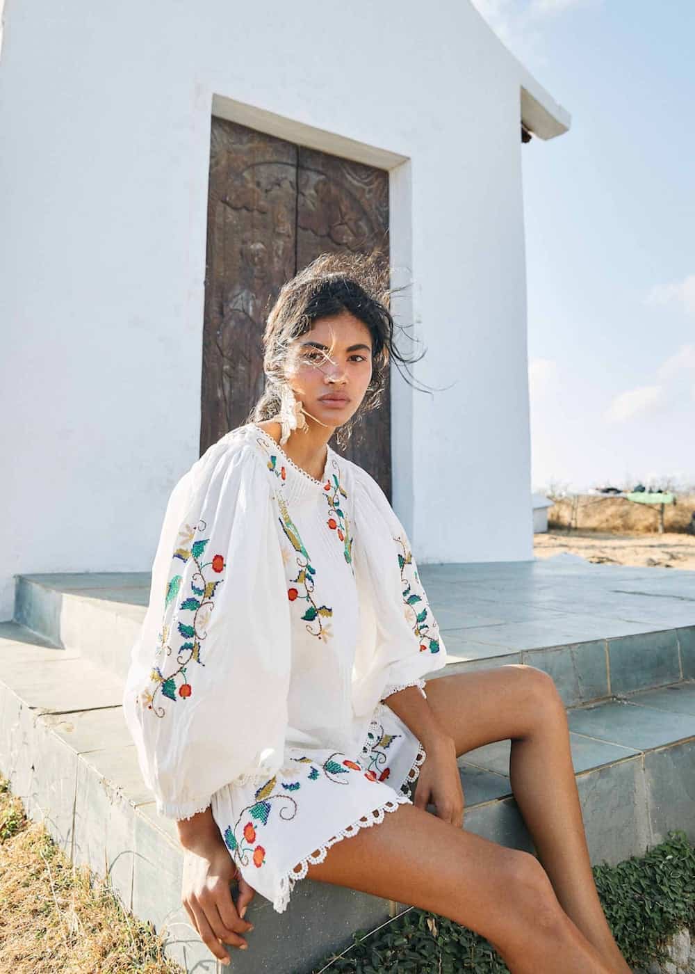 image of a woman sitting in front of a white building wearing an embroidered blouse and shorts set